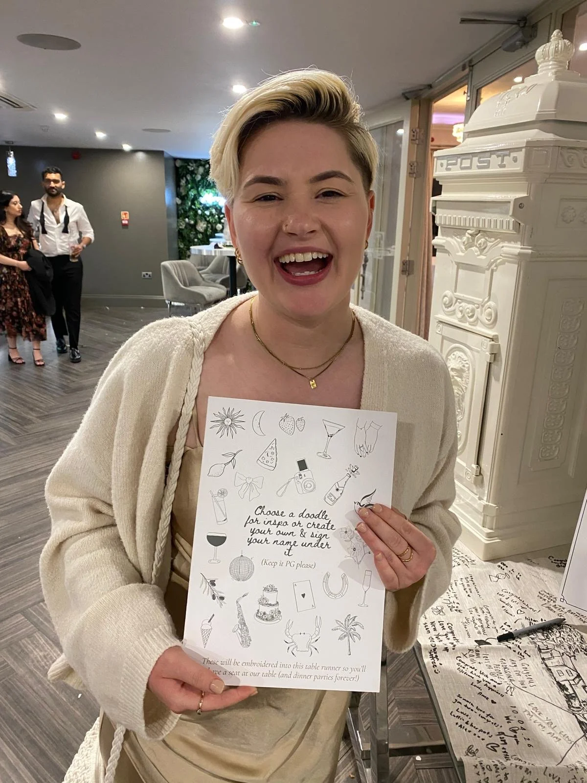 A non-binary person with short blonde hair smiling and holding a sign with wedding-themed doodles at a celebration. There are people in the background and a decorated venue.