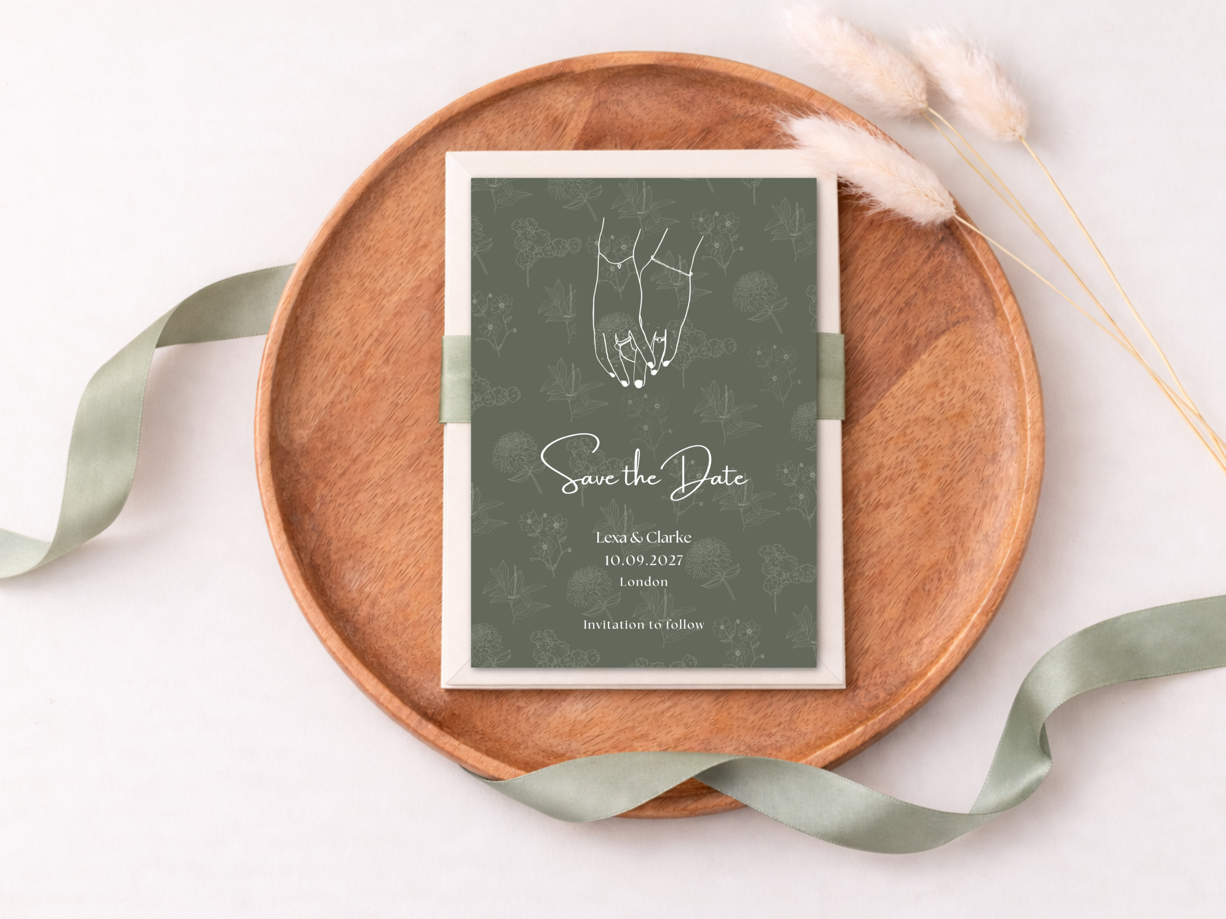A wooden tray holding a dark green wedding invitation with white text and line art, surrounded by light green and beige decorative feathers and ribbons.