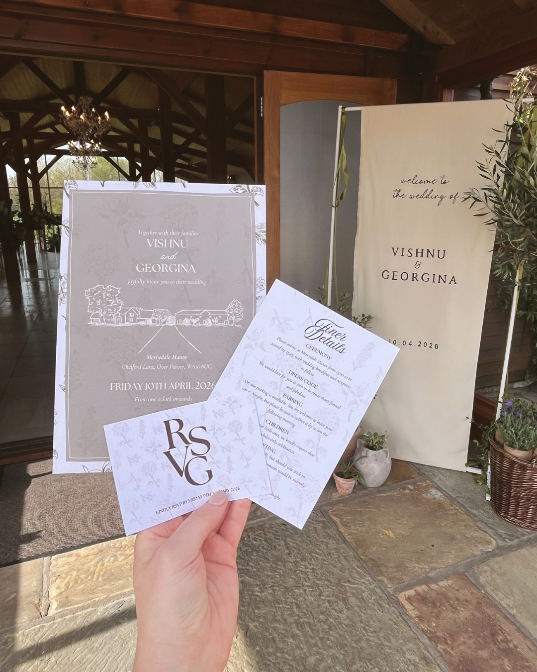 Wedding invitation and RSVP card held in front of an entrance with signs welcoming guests to the wedding of Vishnu and Georgina, April 10, 2026, with potted plants nearby.