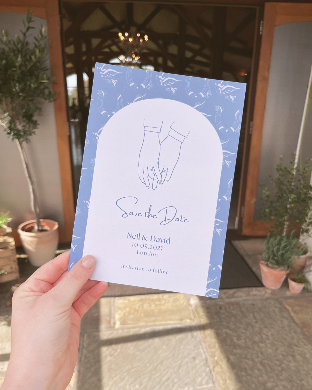 Save the date card with an illustration of two masculine hands holding, for Neil and David's wedding in London on October 9, 2027, featuring a blue background with white decorative elements.