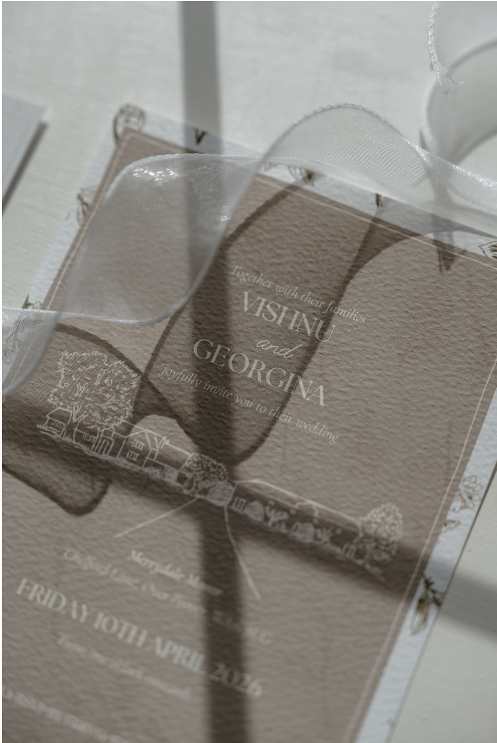 Wedding invitation card with a white ribbon and shadow cast on it, featuring text inviting guests to the wedding of Vishnu and Georgina.