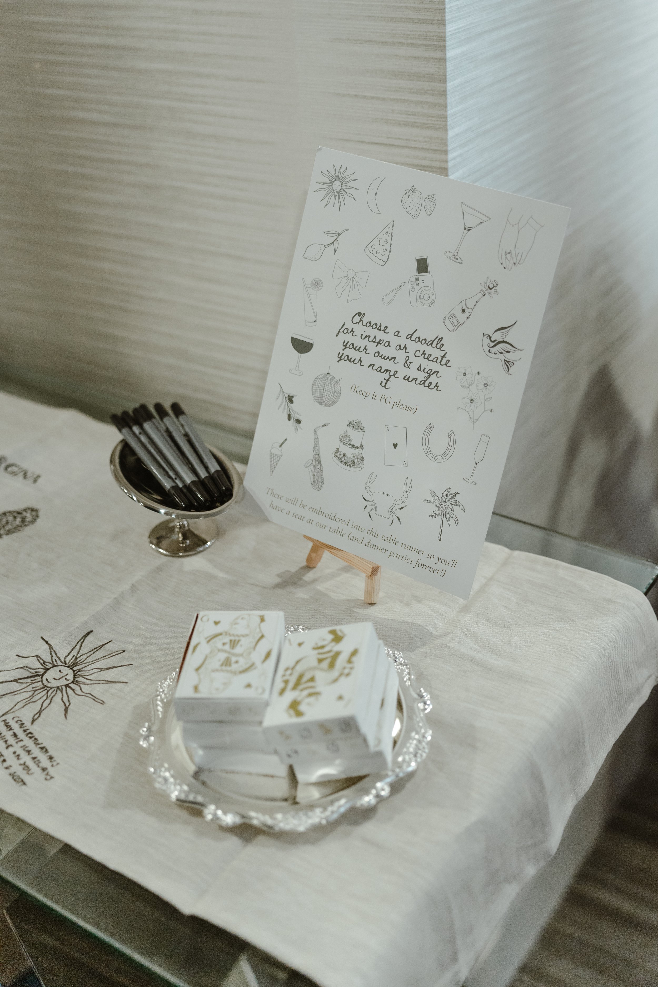 A table with a white tablecloth holding a silver tray with white and gold boxes and a small stack of blocks, and a sign with various doodles and text in black, including a sun, moon, strawberries, cheese, cocktail glass, camera, wine glass, and a mes