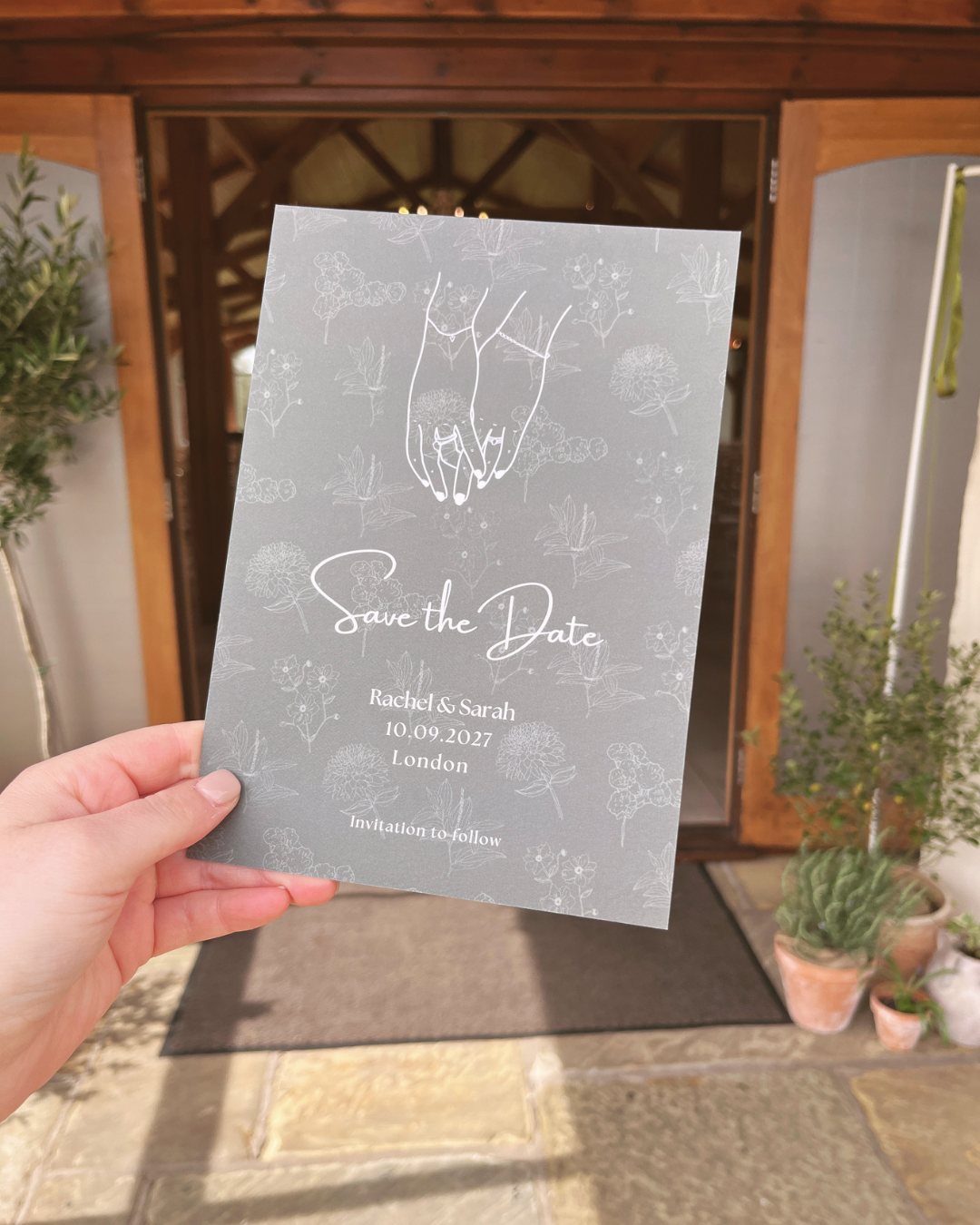 A person holding a 'Save the Date' card with floral designs and an illustration of hands holding each other, outside a wooden building with potted plants nearby.