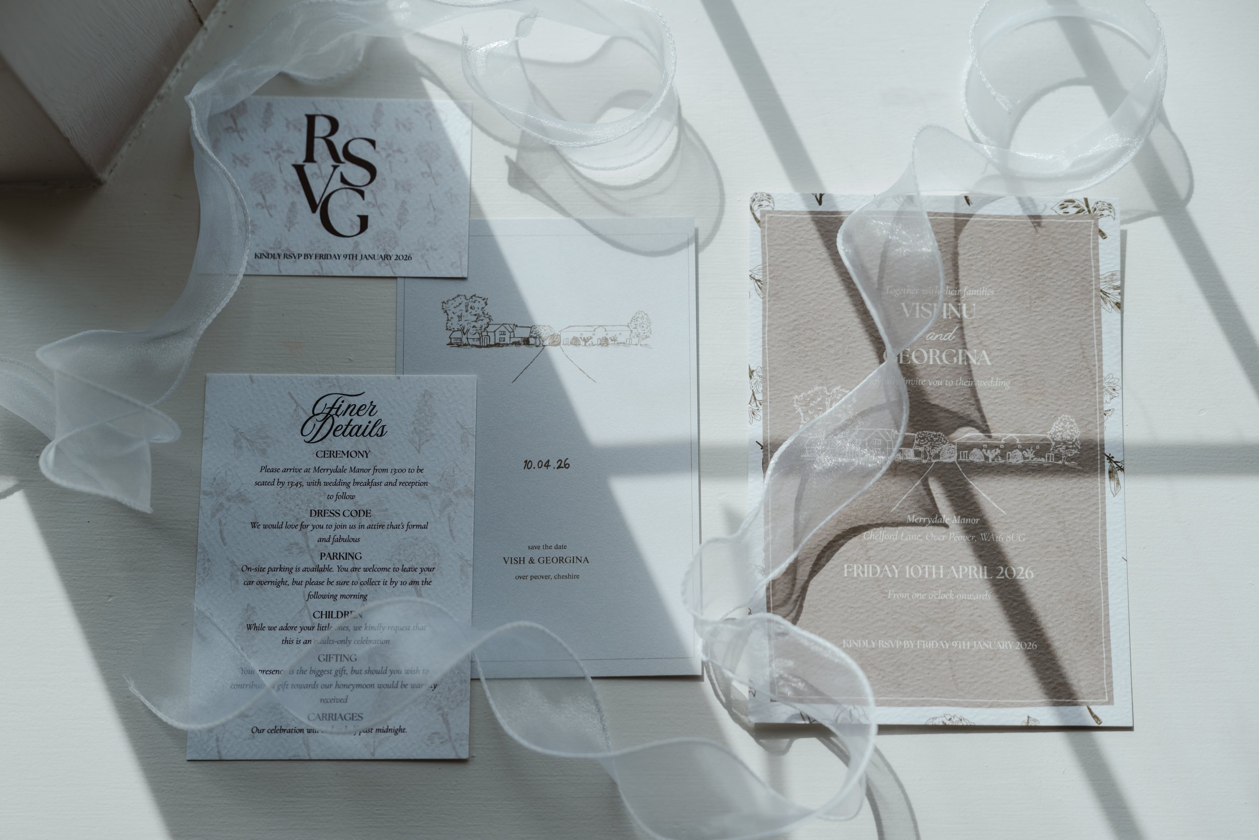 Wedding invitation cards, one with the text 'RSVG' and details about the wedding of Vish and Georgina at Merrydale Manor, with delicate white ribbon flower decorations casting shadows on the cards.