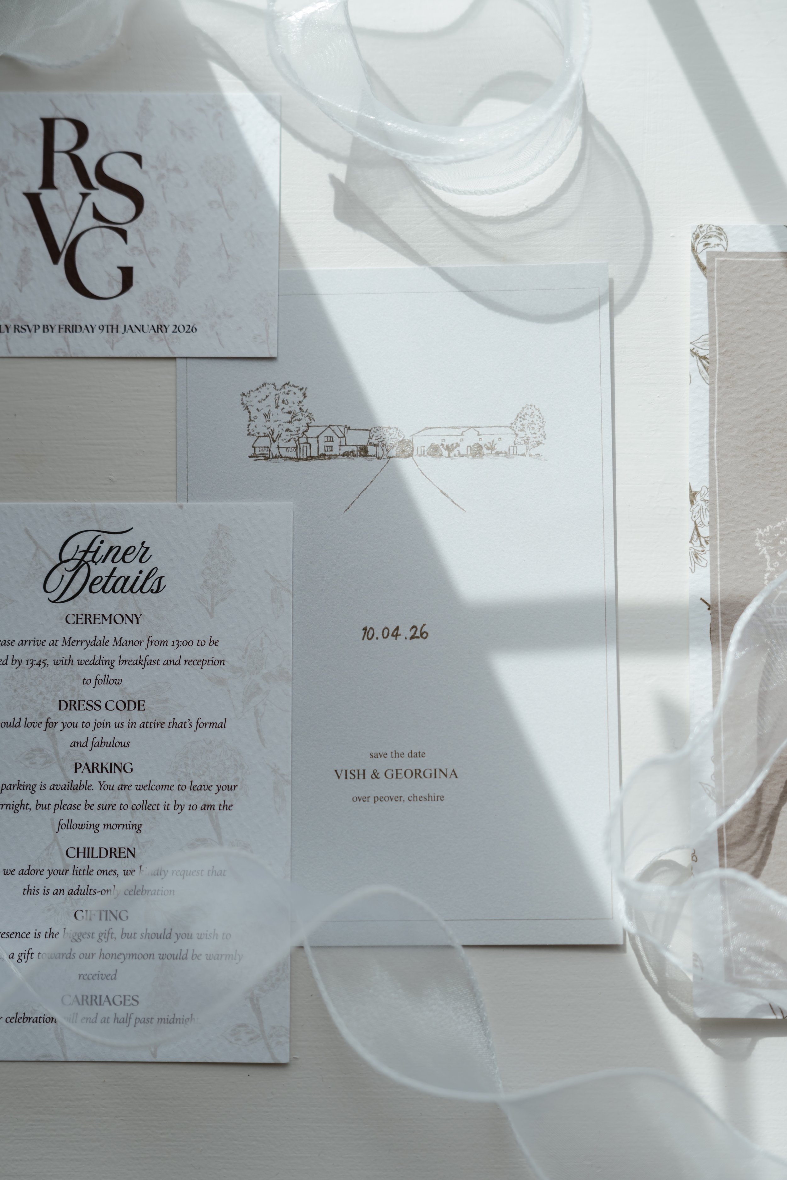 Wedding invitation with illustration of a rural street scene, surrounded by white and beige papers, and a sheer white fabric with shadow cast across the papers.