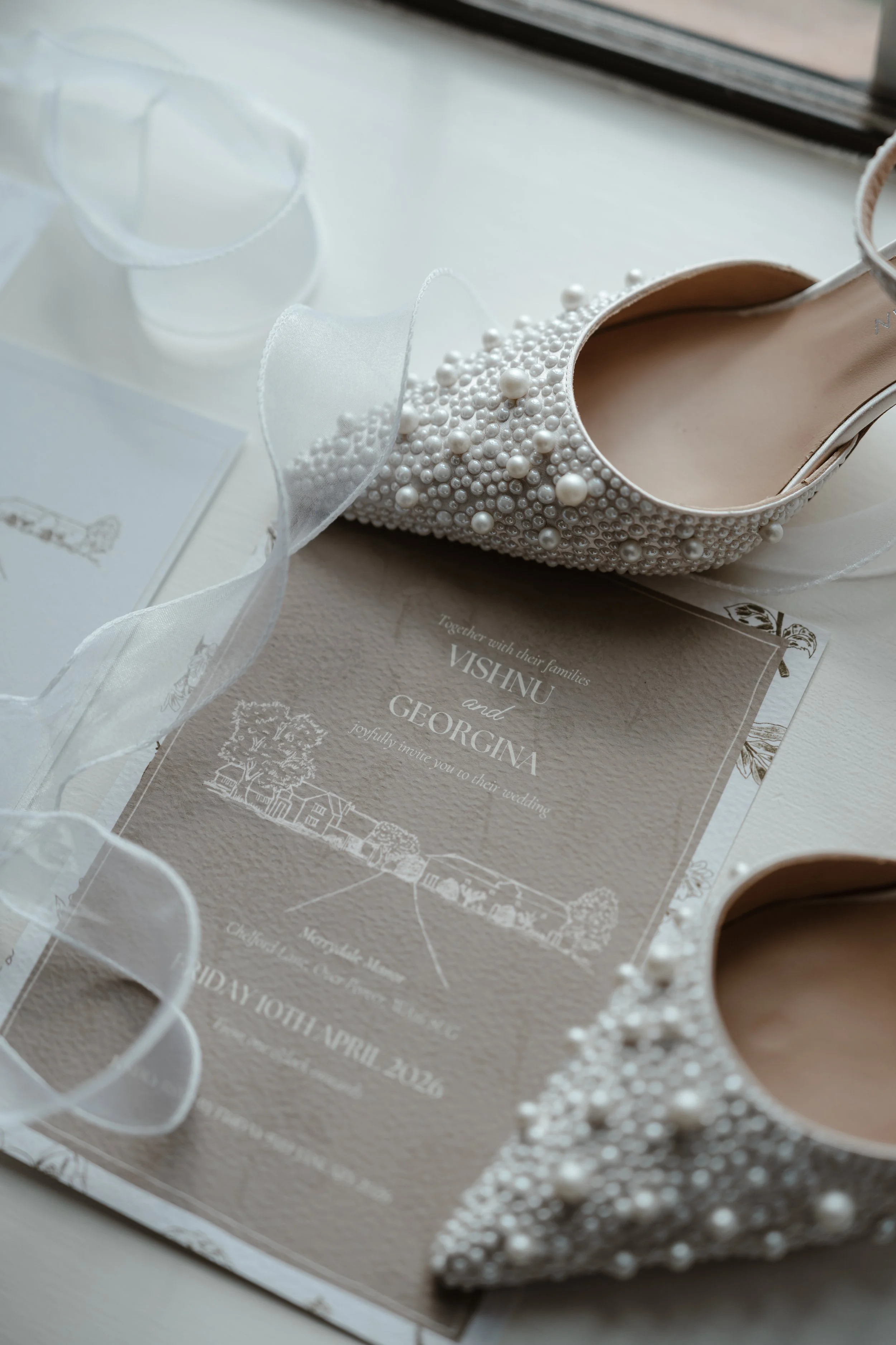 Details of a wedding invitation, a pearl embellished shoe, a card, and a ribbon on a table.