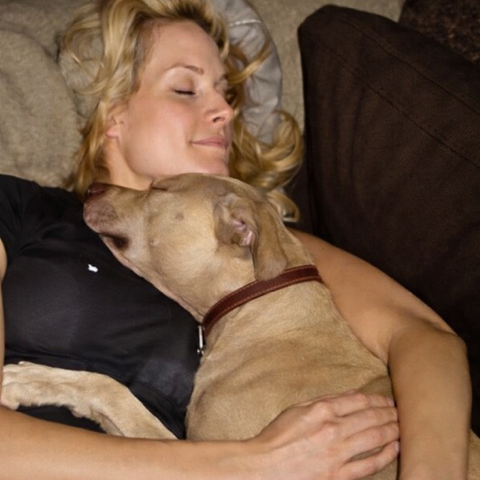 A woman with blonde hair lying on a couch, cuddling with a tan dog with a brown collar, both appearing relaxed and content.