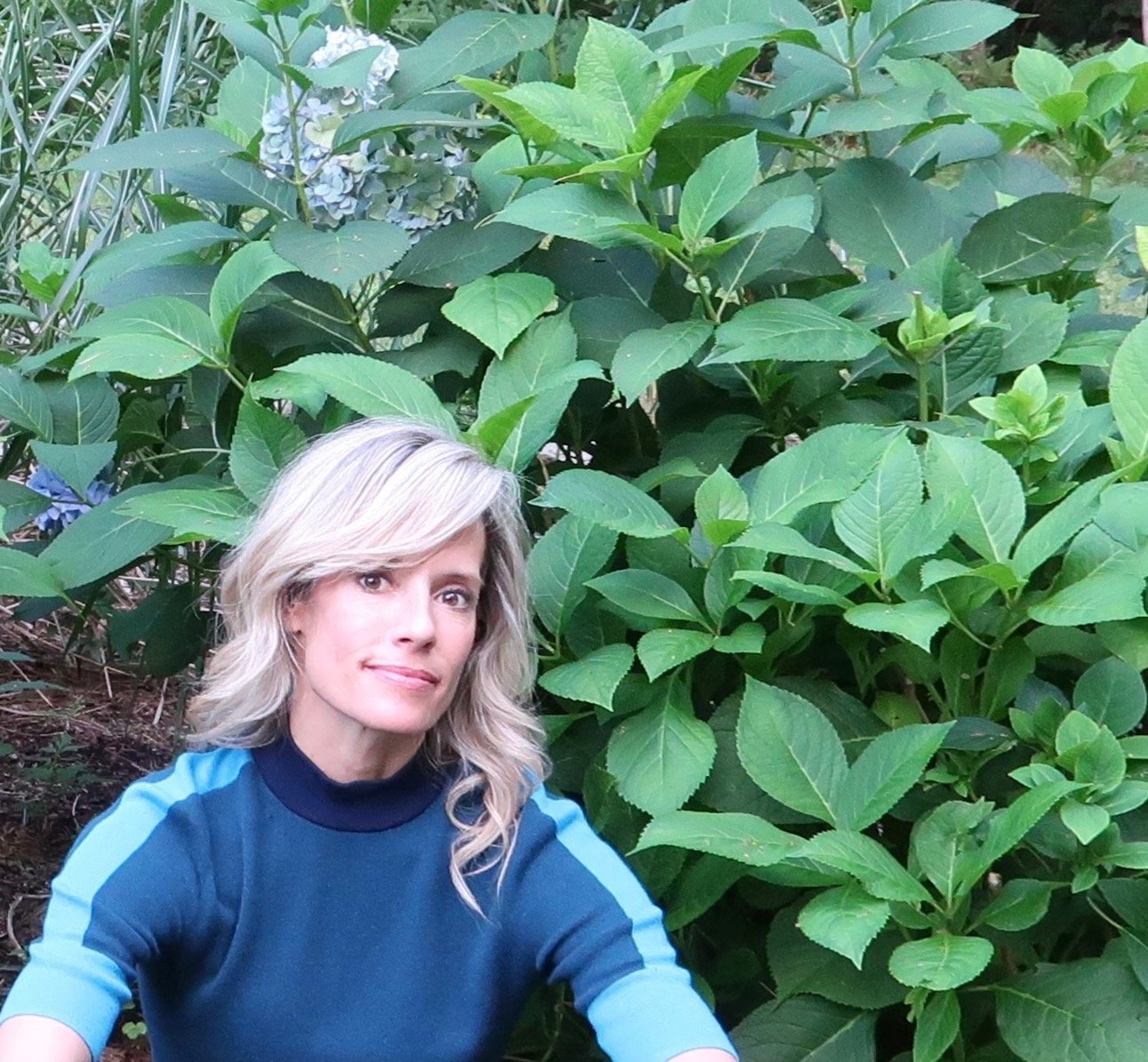 A woman with blonde hair and a blue top taking a selfie in front of lush green bushes and plants in a garden.