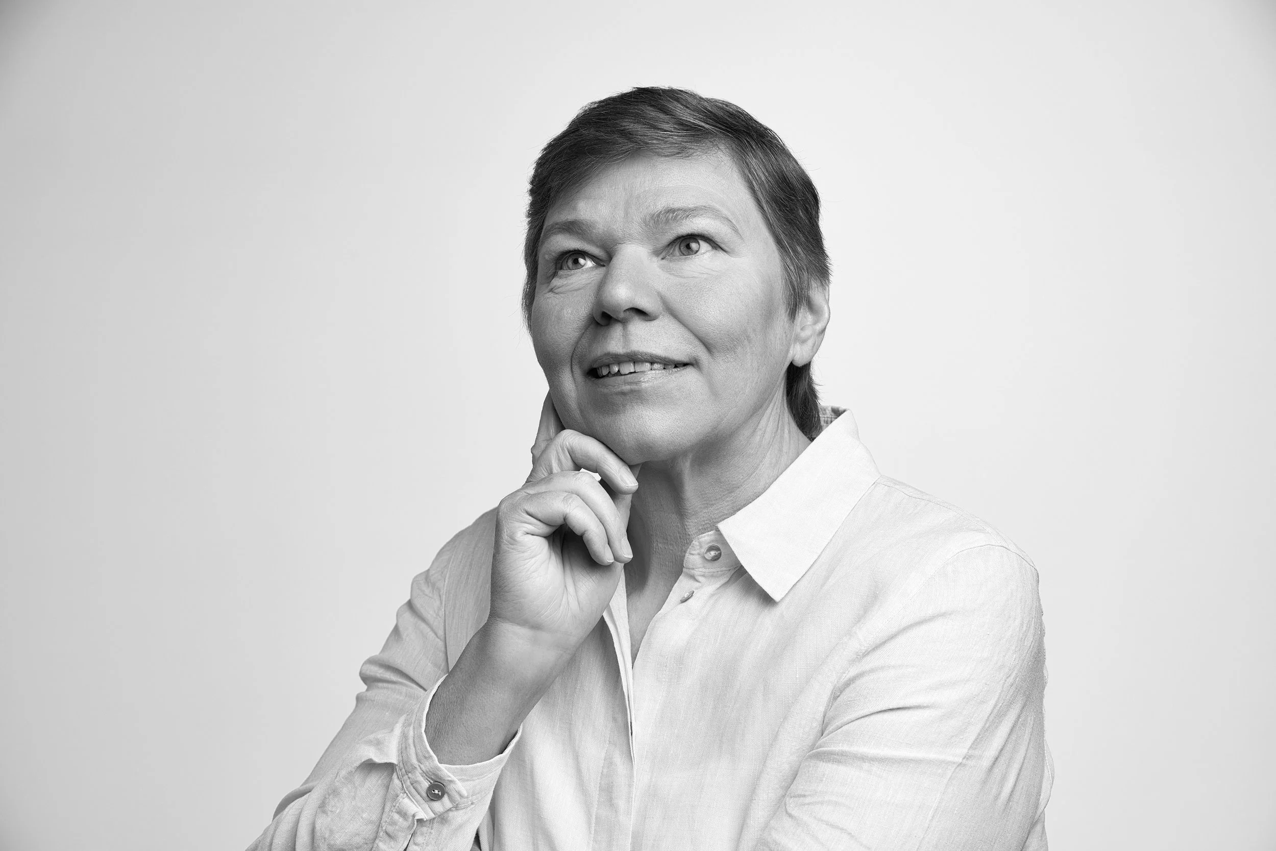 Schwarz-Weiß-Fotografie einer nachdenklichen älteren Frau mit kurzen Haaren, die an ihr Kinn greift und in die Kamera blickt.