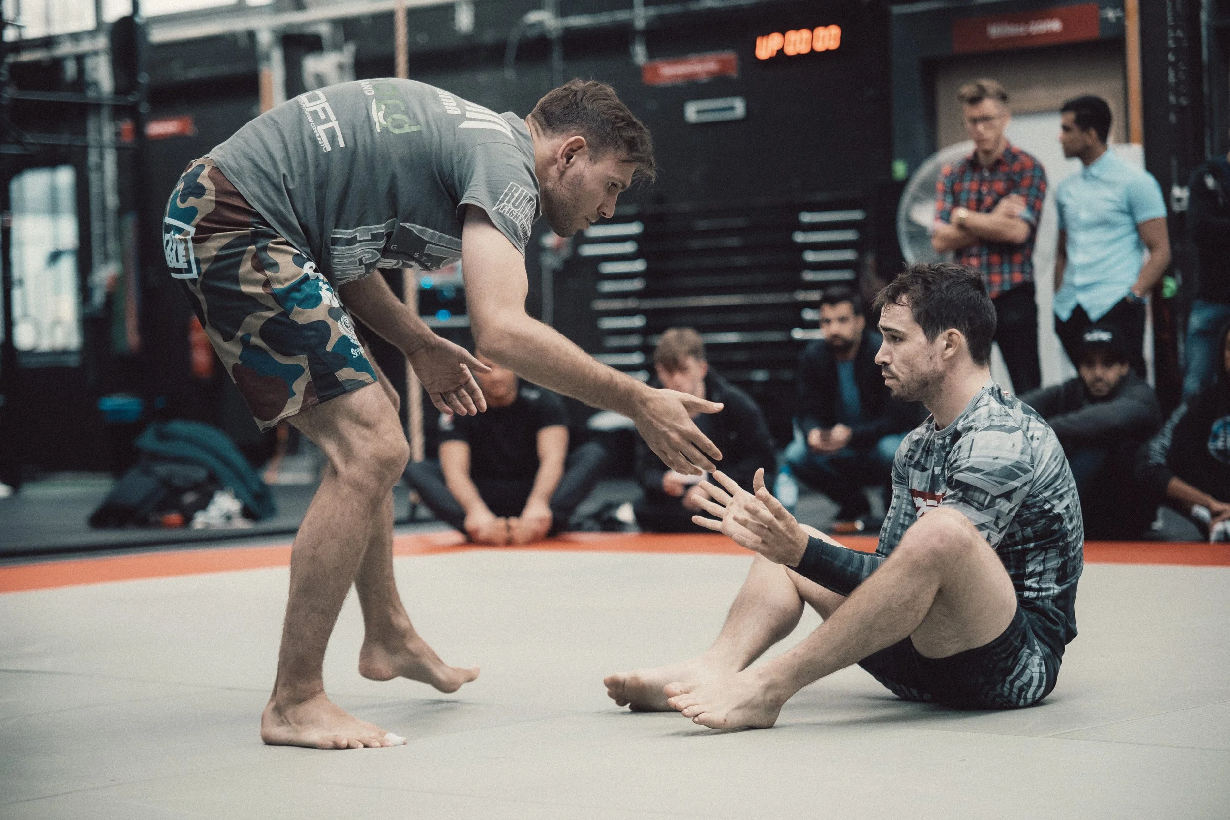 Two men practicing Brazilian Jiu-Jitsu on a mat, with one standing and the other sitting with an arm extended for a handshake, while several people observe in the background.