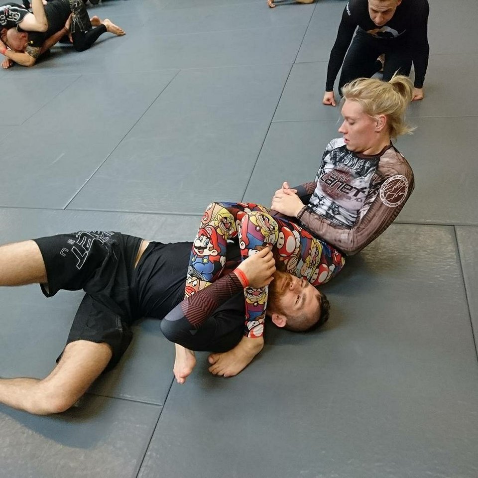 Two mixed martial artists practicing grappling on a padded mat, with one person in a dominant position controlling the other, while additional people watch in the background.