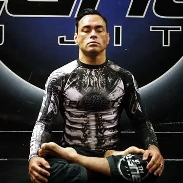 A man sitting cross-legged on the floor in front of a dark backdrop, wearing a black and gray patterned shirt with a graphic design, and holding a small object in his right hand.