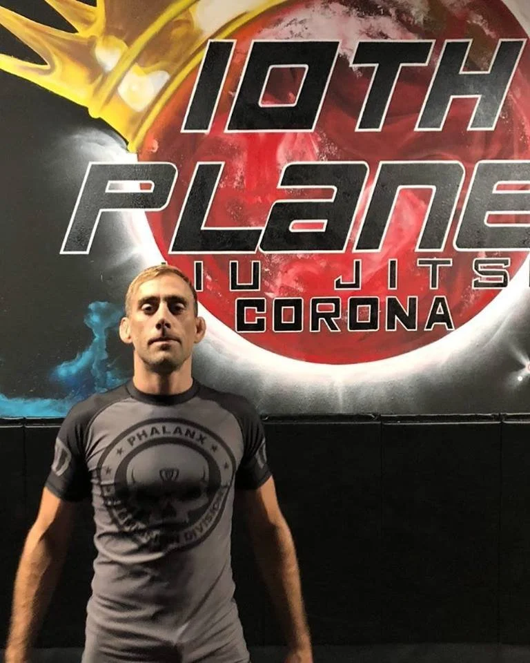 A man standing in front of a poster that says '10th Planet Ju Jitsu Corona'. The poster features a red planet and a yellow robotic hand reaching from the top left.