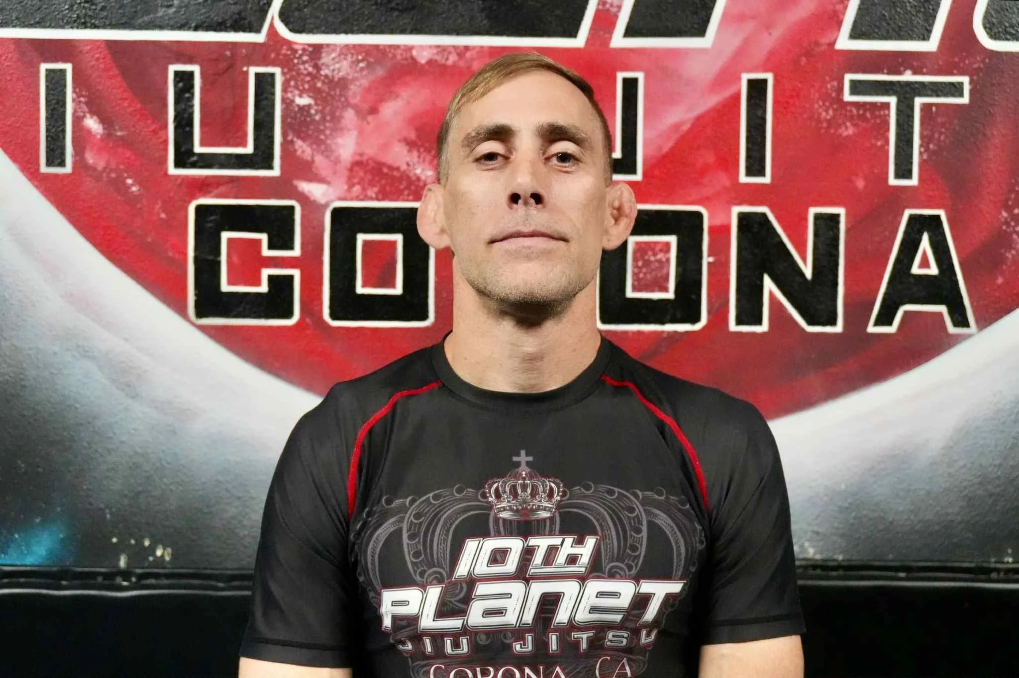A man wearing a black T-shirt with red accents and the text '10th Planet Jiu Jitsu Corona CA'. He stands in front of a large red and black sign that reads 'UFC IT CO CORONA'.