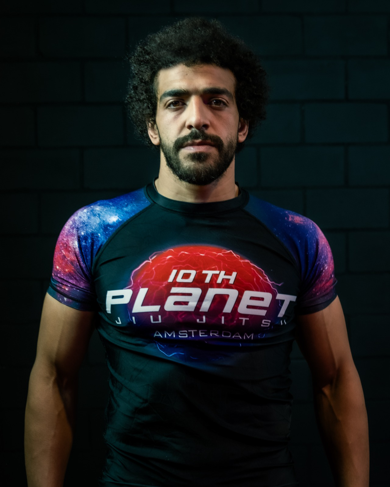 Man with dark curly hair and beard wearing a black T-shirt with cosmic design and the words '10th Planet Jiu Jitsu' and 'Amsterdam' displayed prominently.