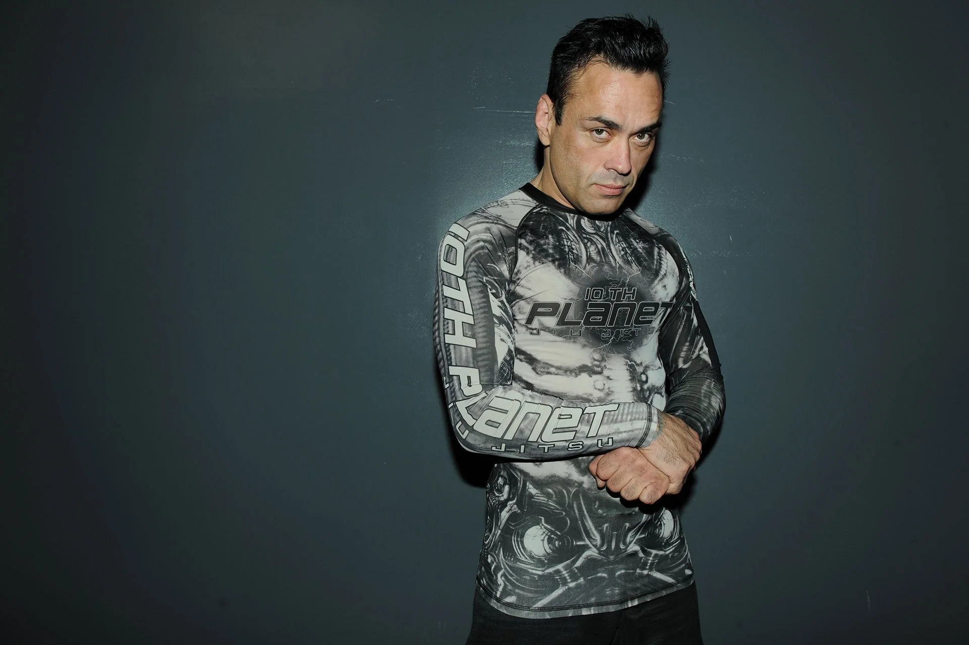 A man with dark hair styled back, wearing a fitted black and gray graphic long-sleeve shirt with the words '10th Planet' on the front and on the sleeve, standing against a dark wall with a confident expression.
