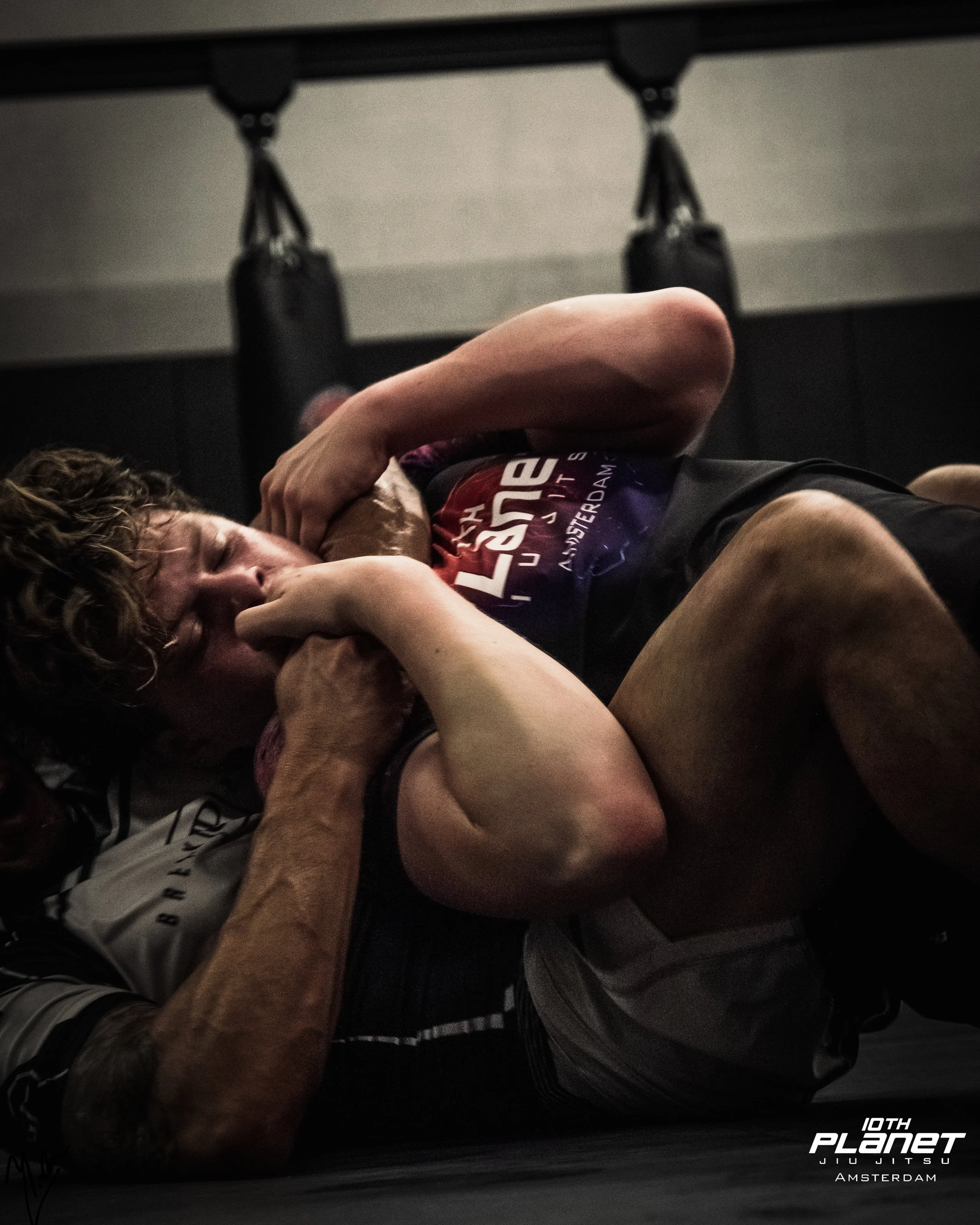Two men grappling in a Brazilian Jiu Jitsu match, with one trying to choke the other from dominant side control in a gym with punching bags in the background.