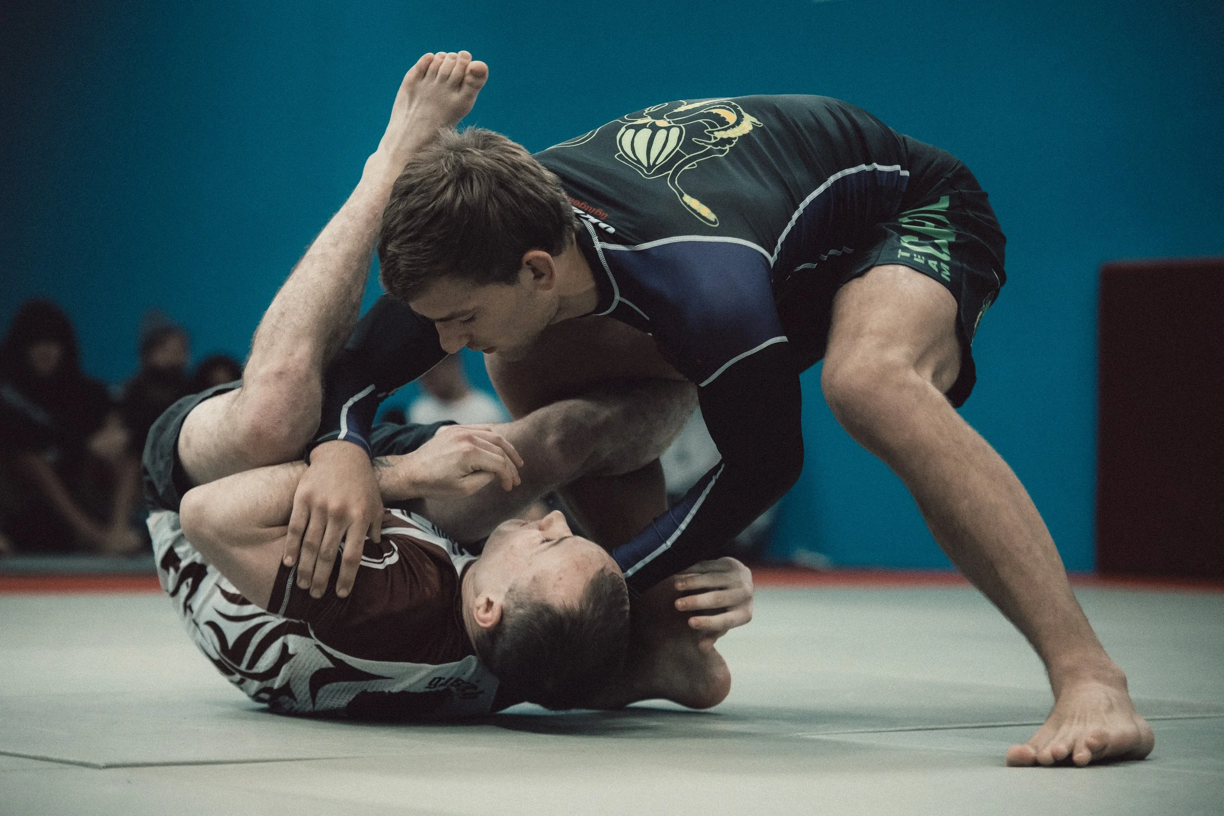 Two men grappling on a gray mat in a Brazilian Jiu-Jitsu match; one man in a black gi with a dragon patch, the other in a maroon and white gi, with spectators in the background.