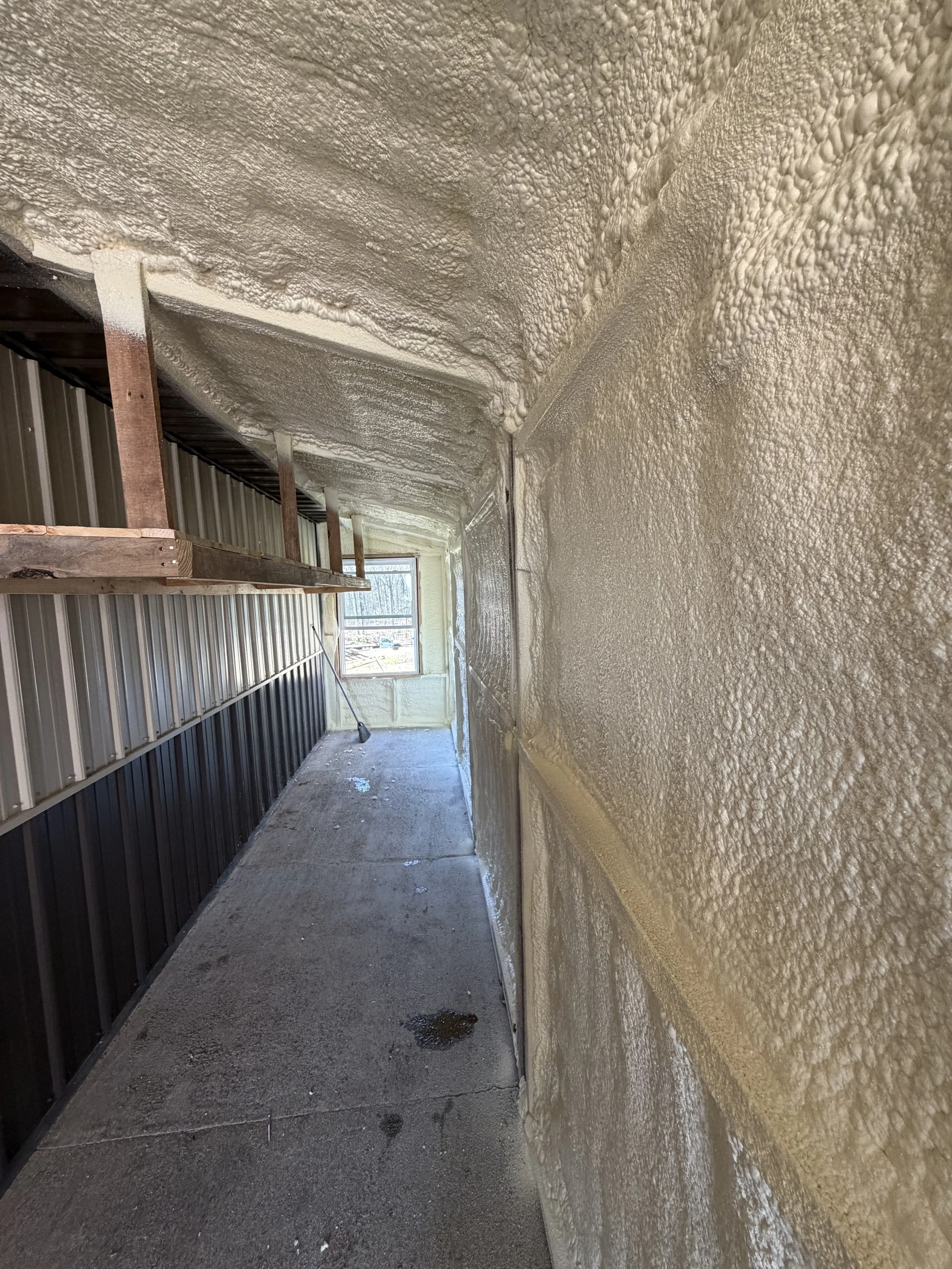 A narrow exterior corridor with foam insulation sprayed onto the walls and ceiling, a concrete floor with water spots, a small window at the end, and a wooden plank structure on the left side.