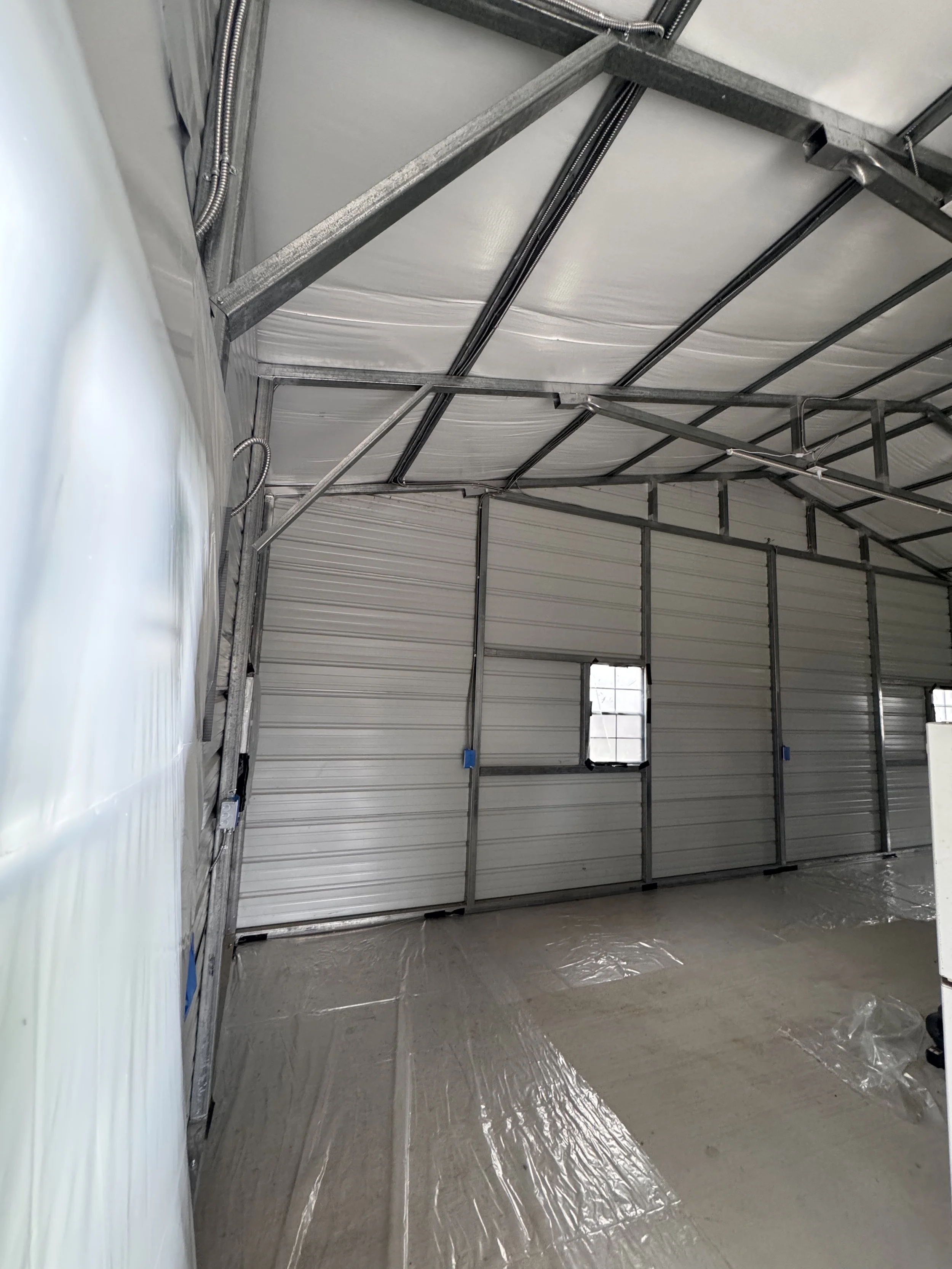 Inside view of a partially constructed garage with metal framing and insulated walls, with two small windows and plastic covering on the floor.