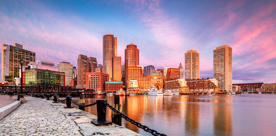City skyline at sunset reflecting on water, with a promenade in the foreground.