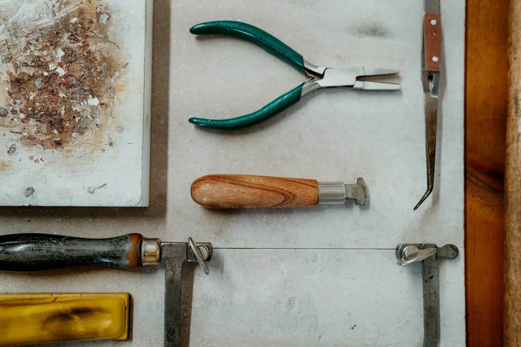 A pair of green-handled pliers, a wooden-handled tool, and part of a saw are arranged on a light-colored work surface.