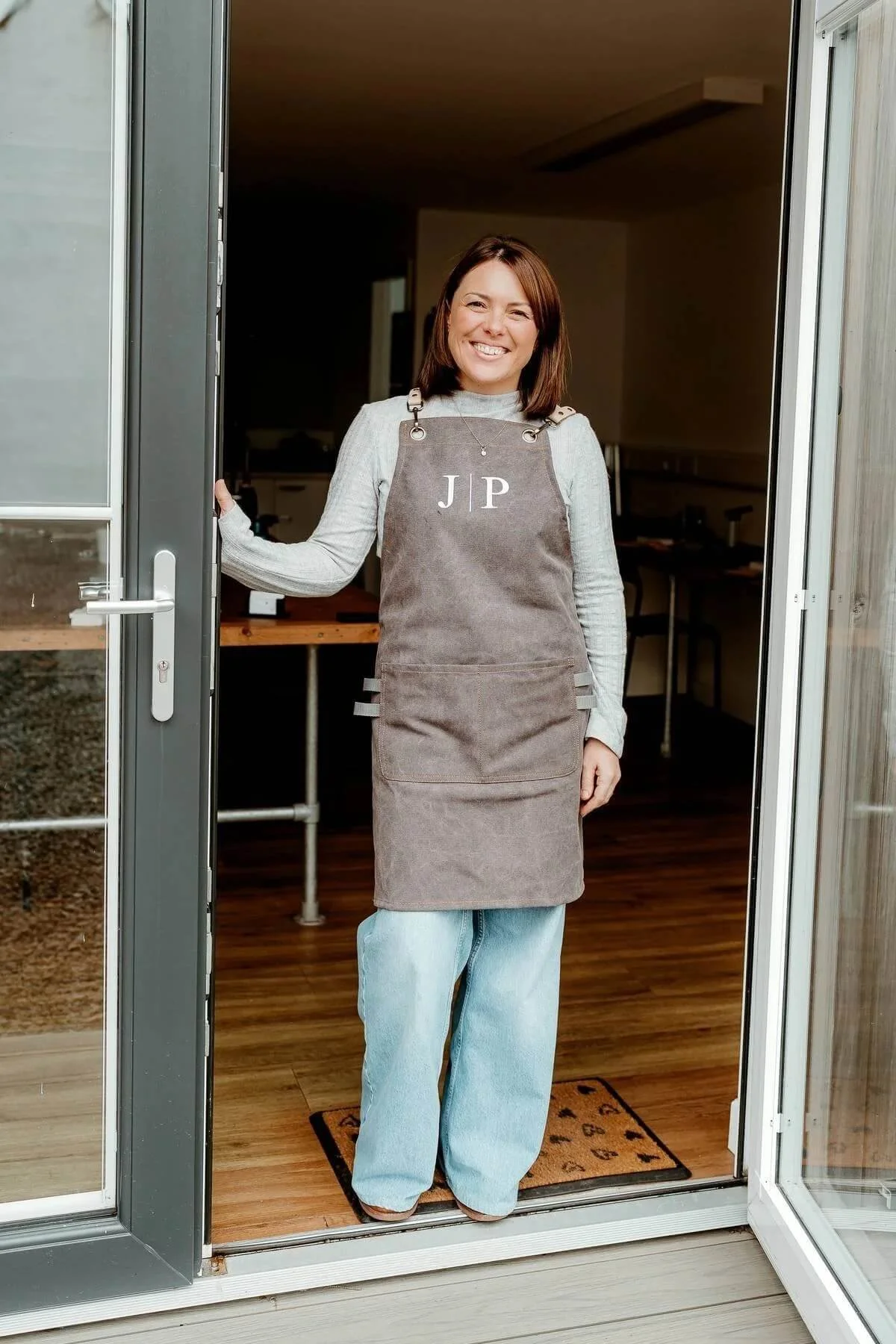 A woman standing in a doorway, smiling, wearing a gray apron with the initials 'J P' embroidered on it and casual clothing underneath, inside a room with wooden flooring.