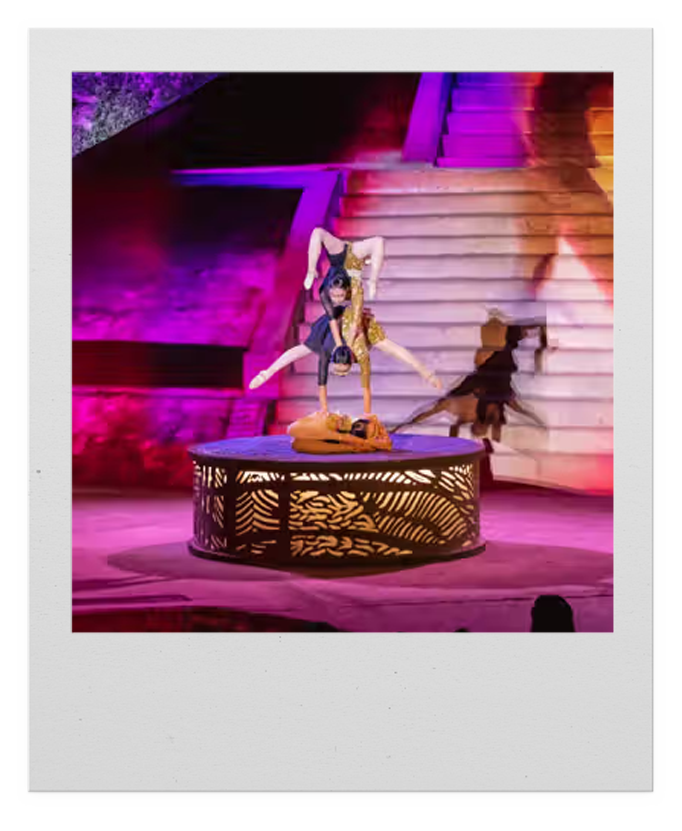 A performer doing an acrobatic handstand on a table during a colorful stage show, with stairs and vibrant lighting in the background.