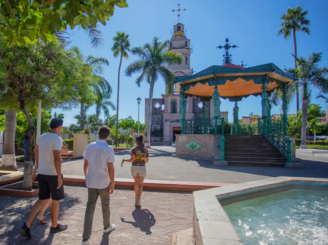 A small plaza with a fountain and a gazebo featuring a statue or monument in the center. Four people are walking toward the monument, surrounded by palm trees and lush greenery, with a bright blue sky overhead.