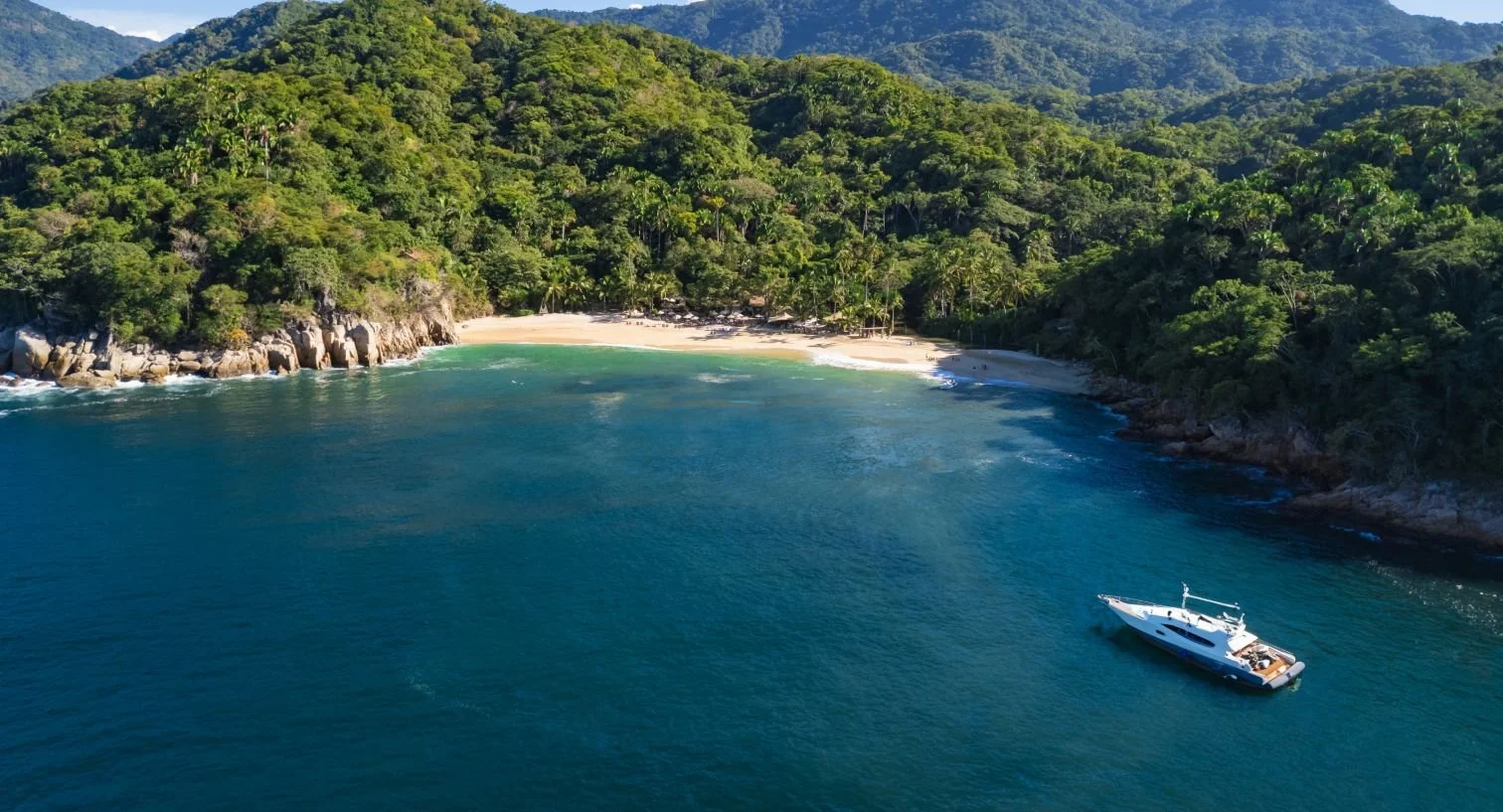 Aerial view of a tropical coastline with a beach, lush green forest, and a yacht in the water.