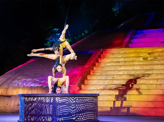 Three acrobats performing a balancing act on a circular platform with stairs and colorful lighting in the background.