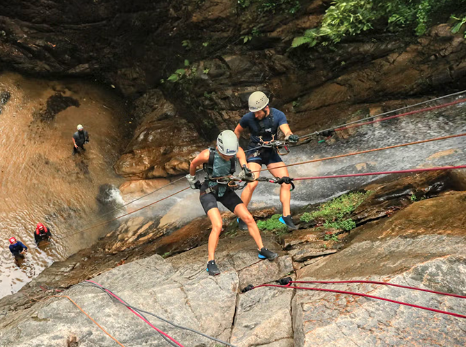 People participating in a canyoning adventure, climbing on wet rocks with safety helmets and harnesses, with a waterfall nearby.