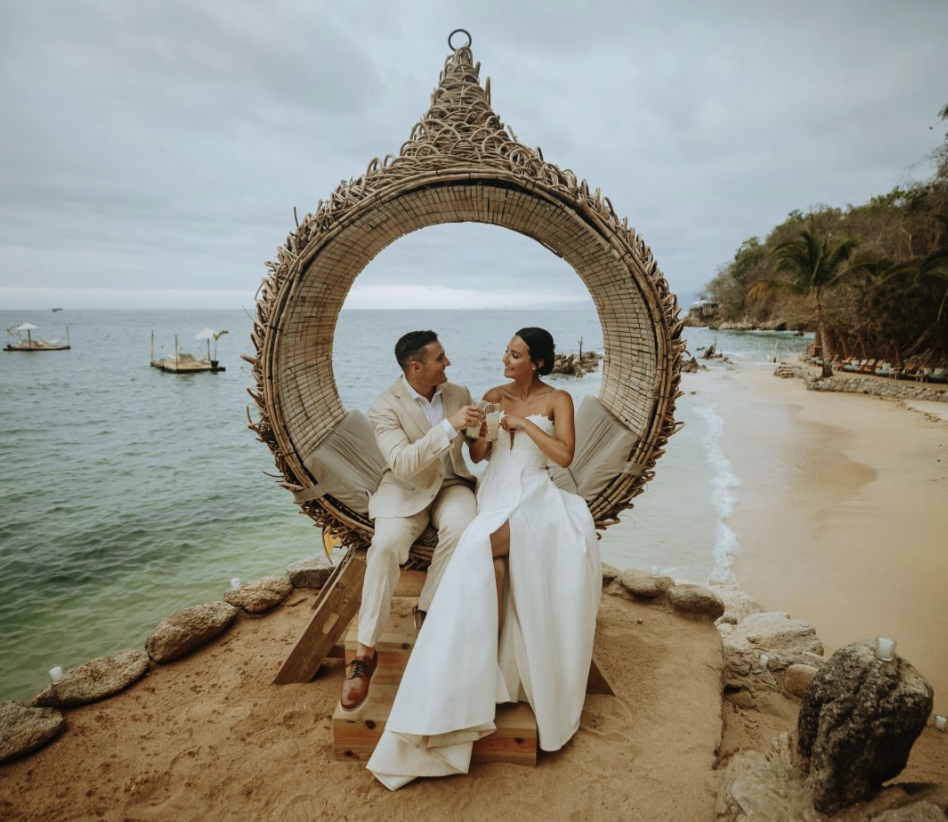Couple in wedding attire sitting in a wicker hanging chair on a beach, holding drinks, with the ocean and boats in the background.