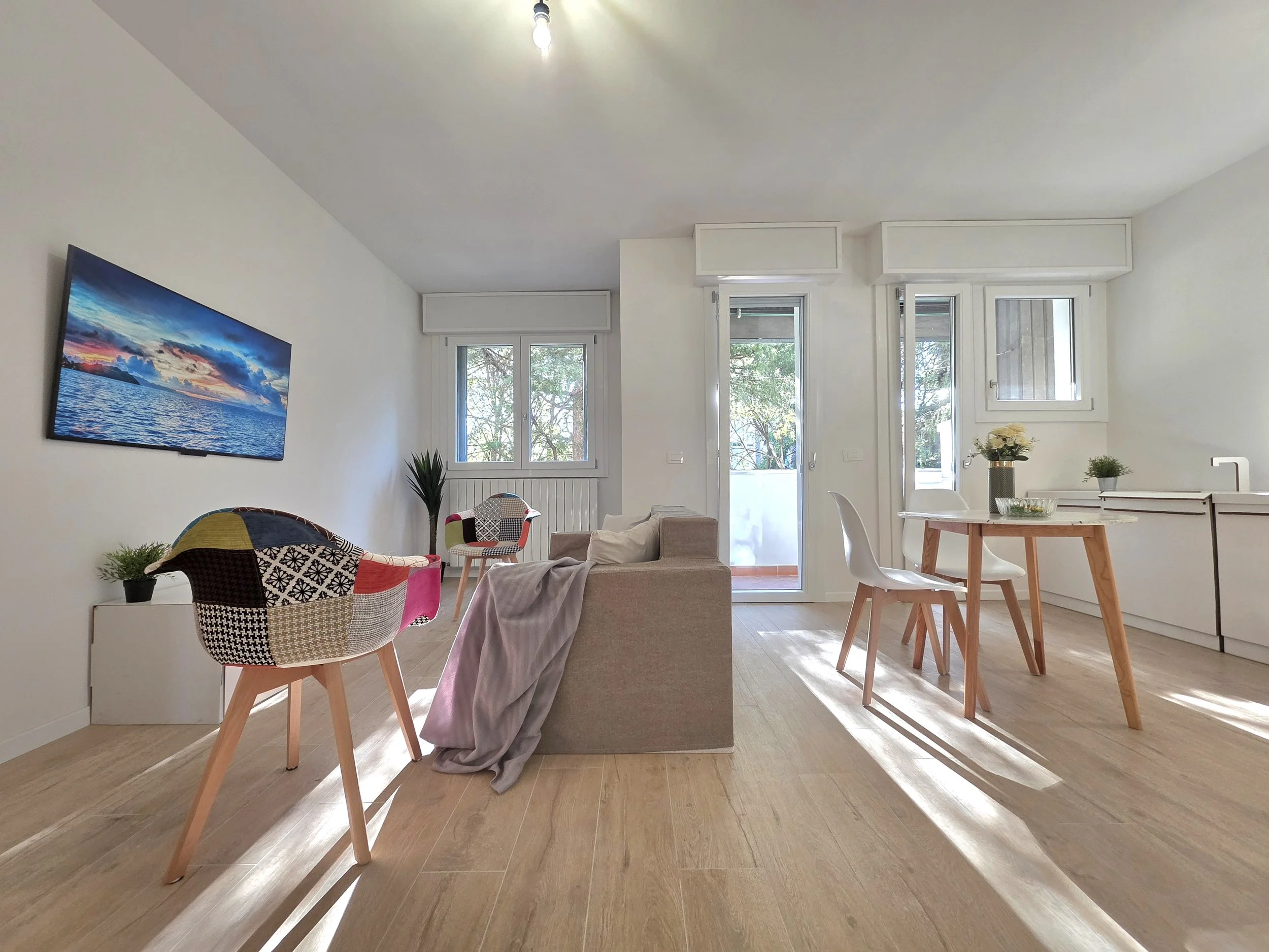 Soggiorno minimalista con divano beige, tavolo rotondo con sedie bianche, pareti bianche, grandi finestre e porta finestra che porta a un balcone, TV a schermo piatto sulla parete, decorazioni floreali e piante.