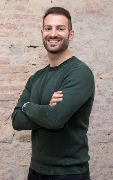 Uomo sorridente con braccia incrociate davanti a un muro di mattoni.