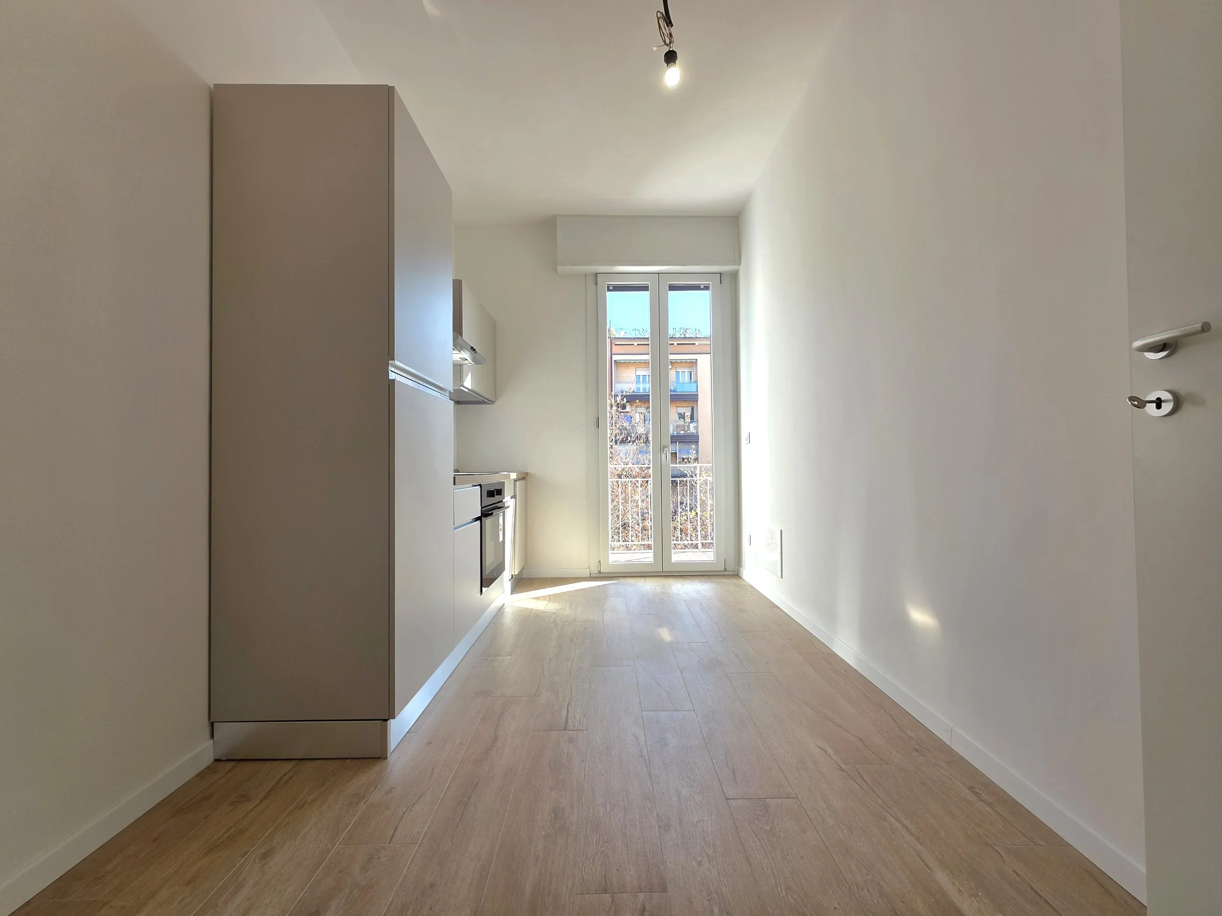 Cucina minimalista con pareti bianche, pavimento di legno chiaro, grande finestra che dà su un balcone con vista su edifici circostanti, cucina moderna con elettrodomestici integrati.
