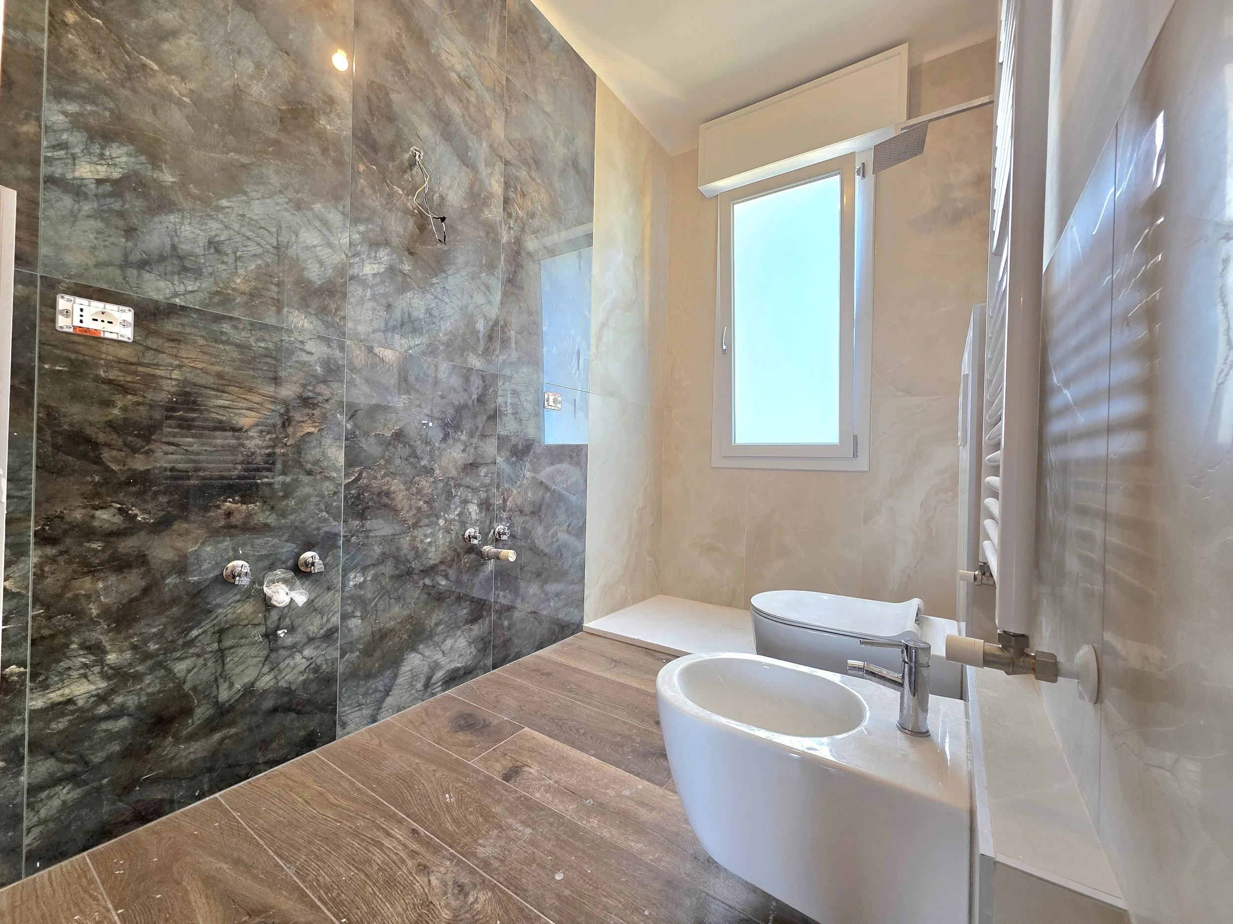 Bagno in fase di completa VERNICIATURA con pareti in marmo scuro e pavimento in legno, finestra aperta, sanitari bianco e radiatore