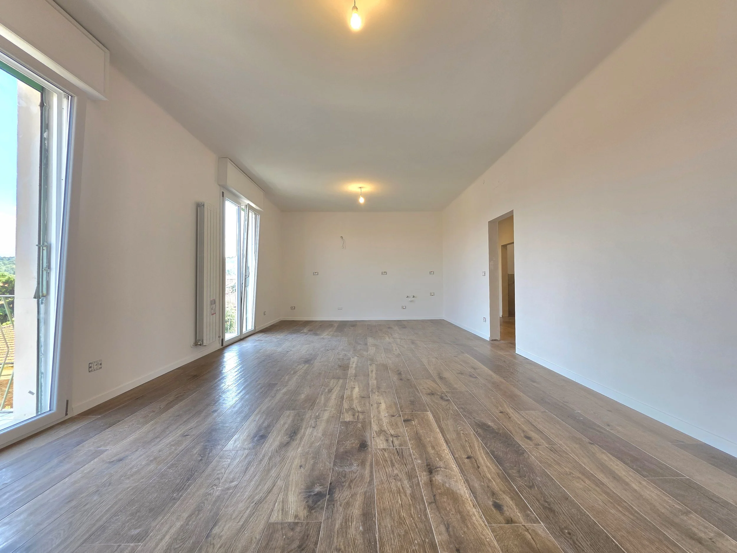 Interno vuoto di una stanza spaziosa con pareti bianche, pavimento in legno e grandi finestre che permettono l'ingresso di molta luce naturale.