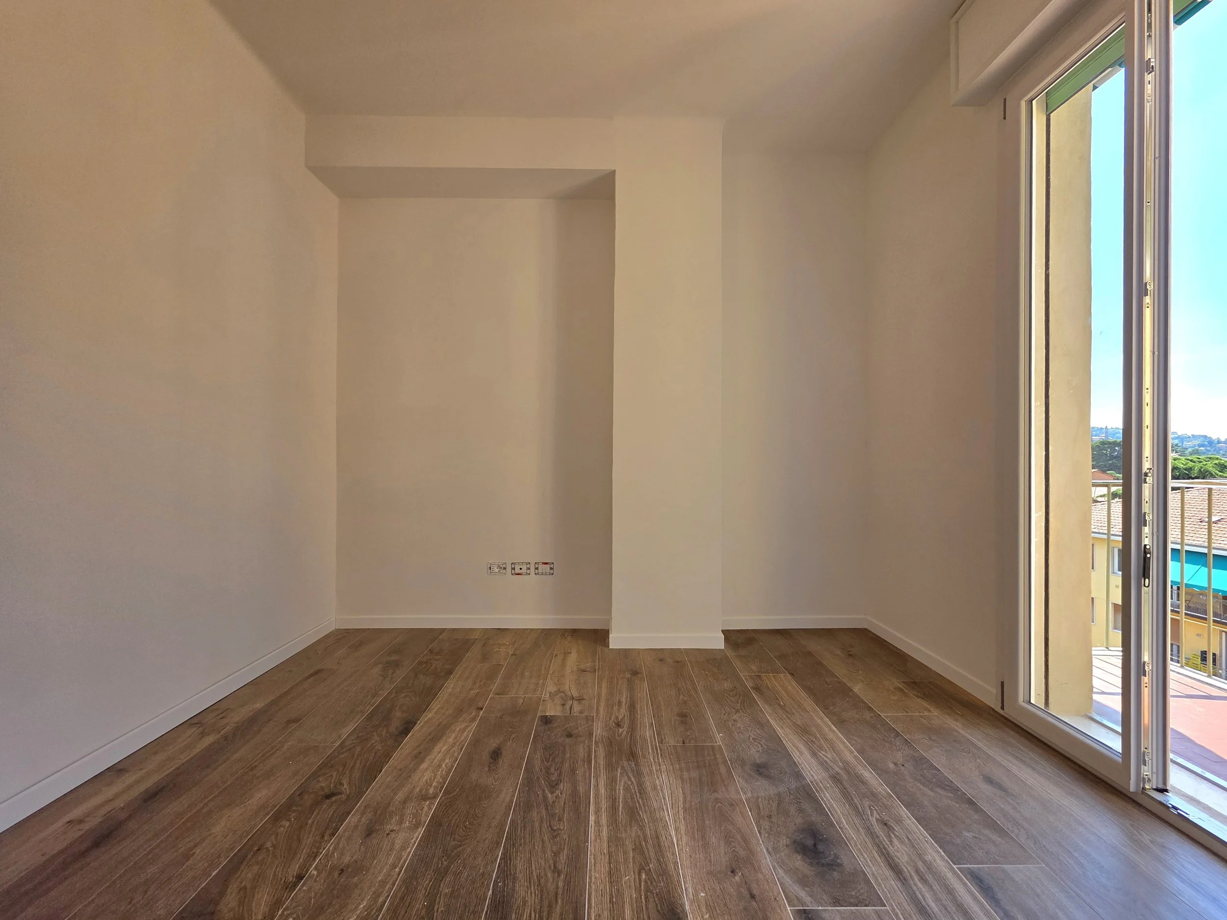 Stanza vuota con pareti bianche, pavimento in legno e grande finestra con balcone che lascia entrare molta luce naturale.
