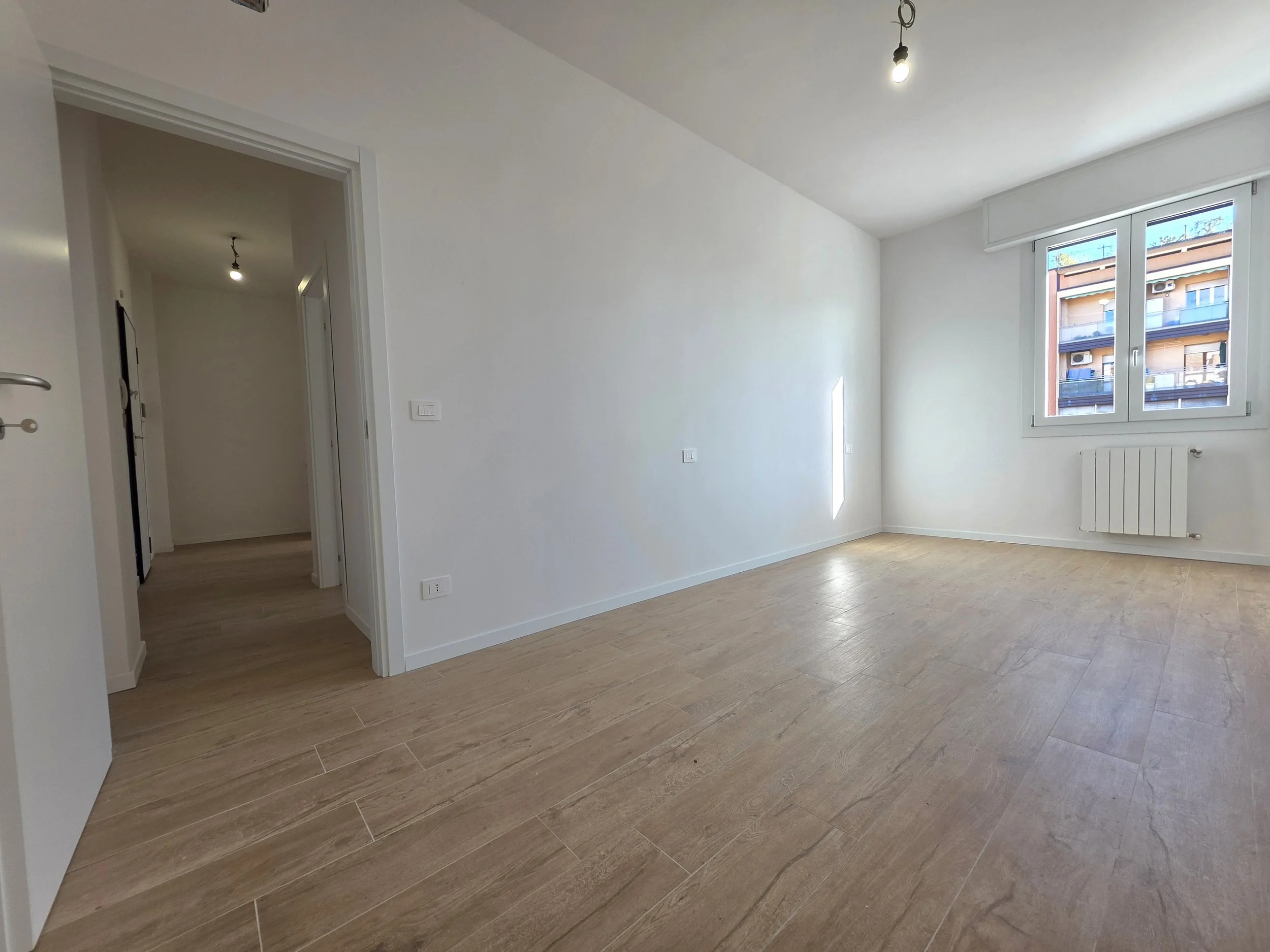 Stanza vuota con pareti bianche, pavimento in legno chiaro, una finestra grande che dà su edifici urbani, e un radiatore sotto la finestra.