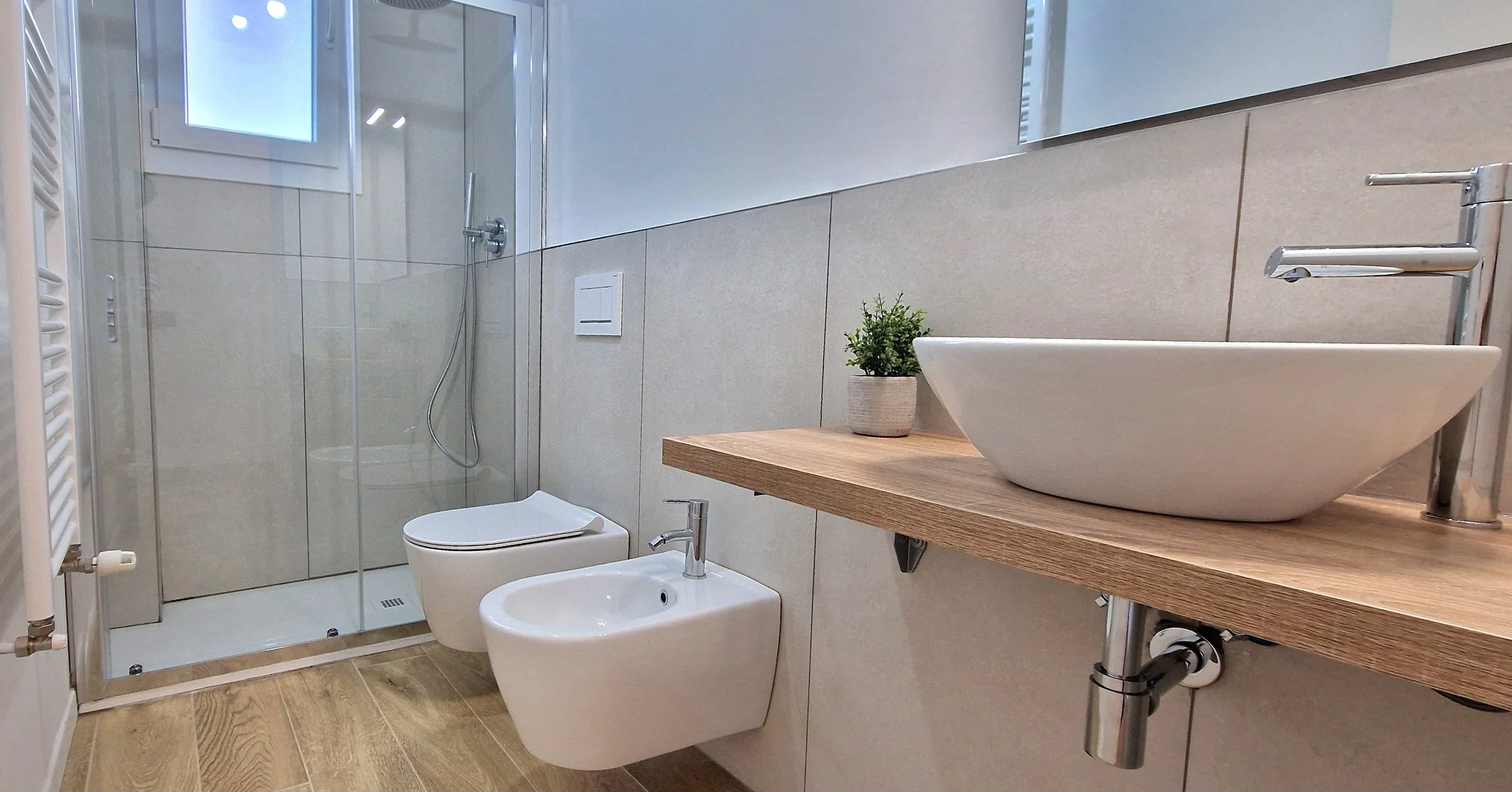 Bagno moderno con doccia, lavabo su mobile in legno, piante e toalette bianca, pareti chiare e pavimento in legno.