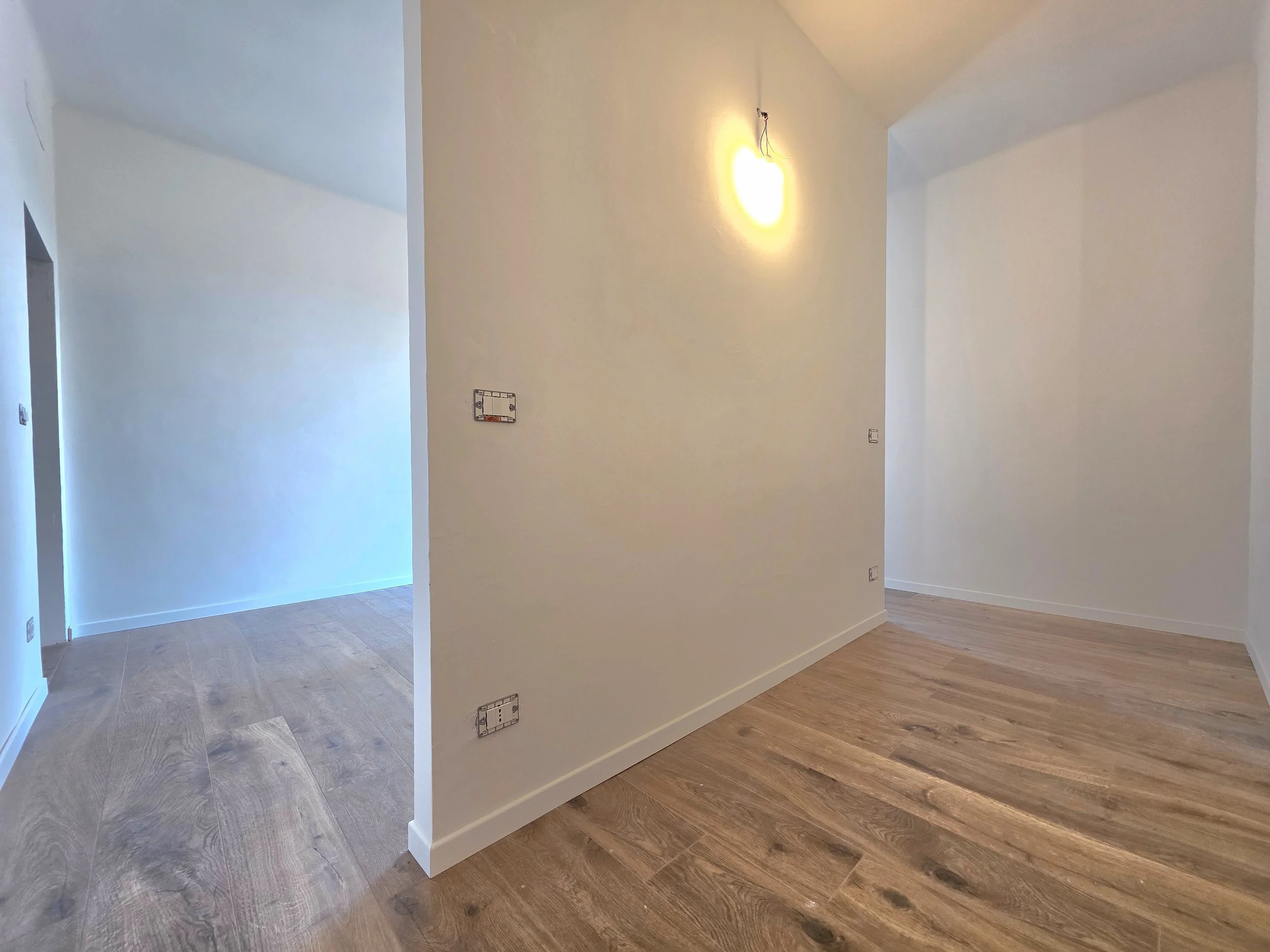 Interno di una stanza in fase di ristrutturazione con pareti bianche, pavimento in legno e una luce accesa sulla parete.