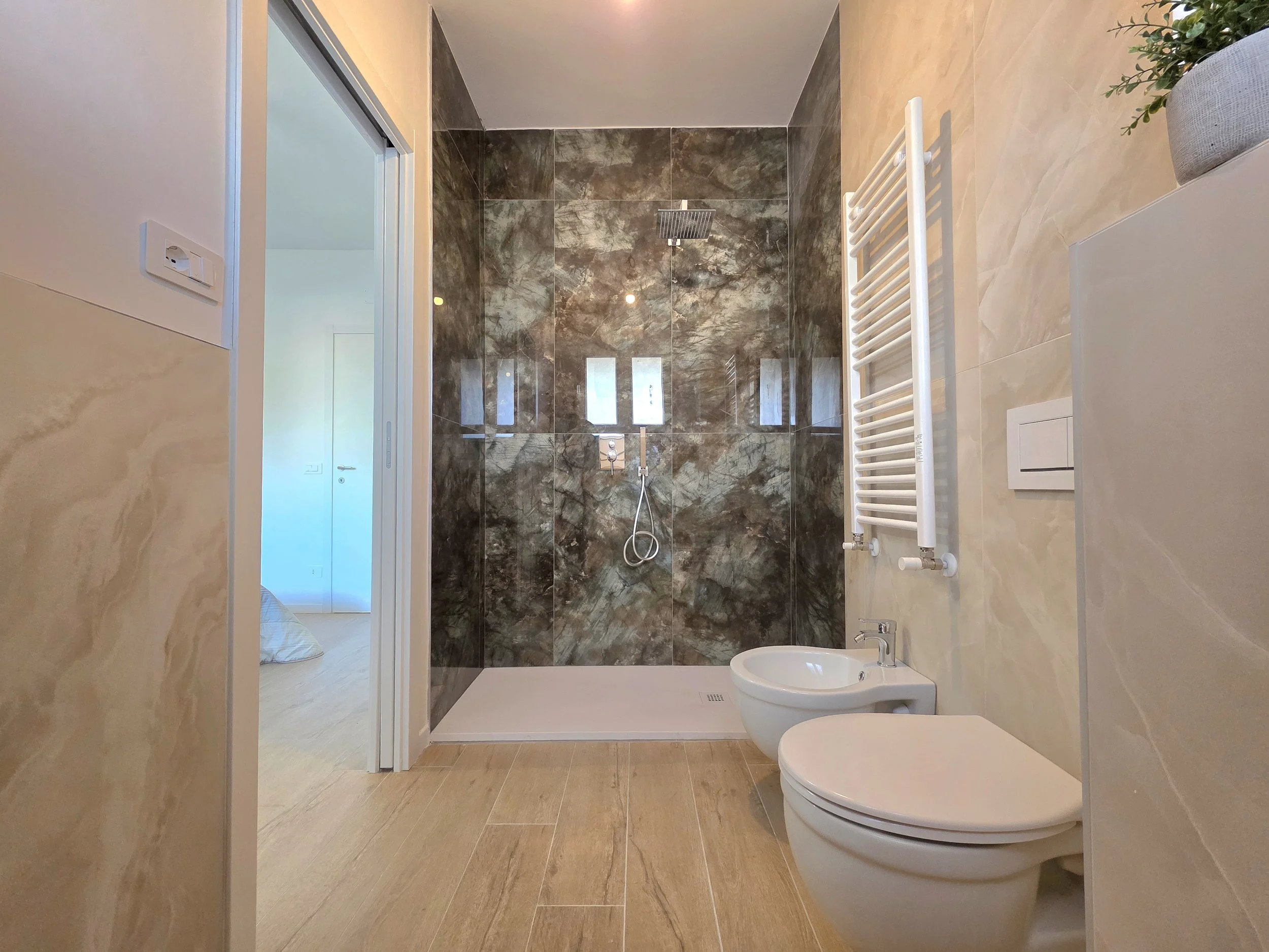 Bagno moderno con pareti in marmo scuro e beige, doccia a pavimento con pareti in marmo scuro, termosifone bianco verticale, bidet e WC bianco, pianta in vaso e pareti beige.