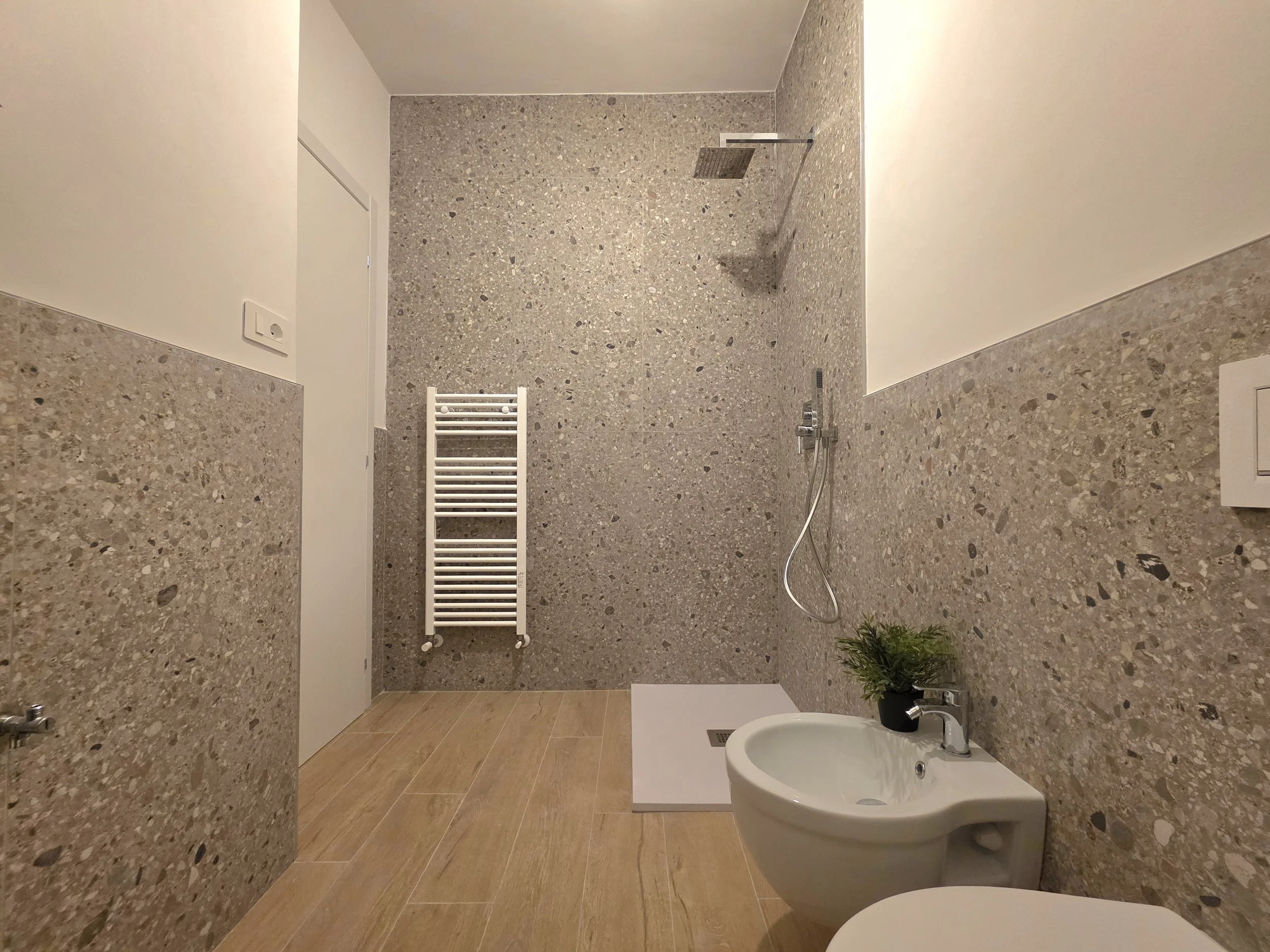 Bagno con pareti rivestite in pietra, doccia a pioggia, termosifone bianco, lavandino con pianta verde, pareti in parte biene e parte bianca, pavimento in piastrelle beige