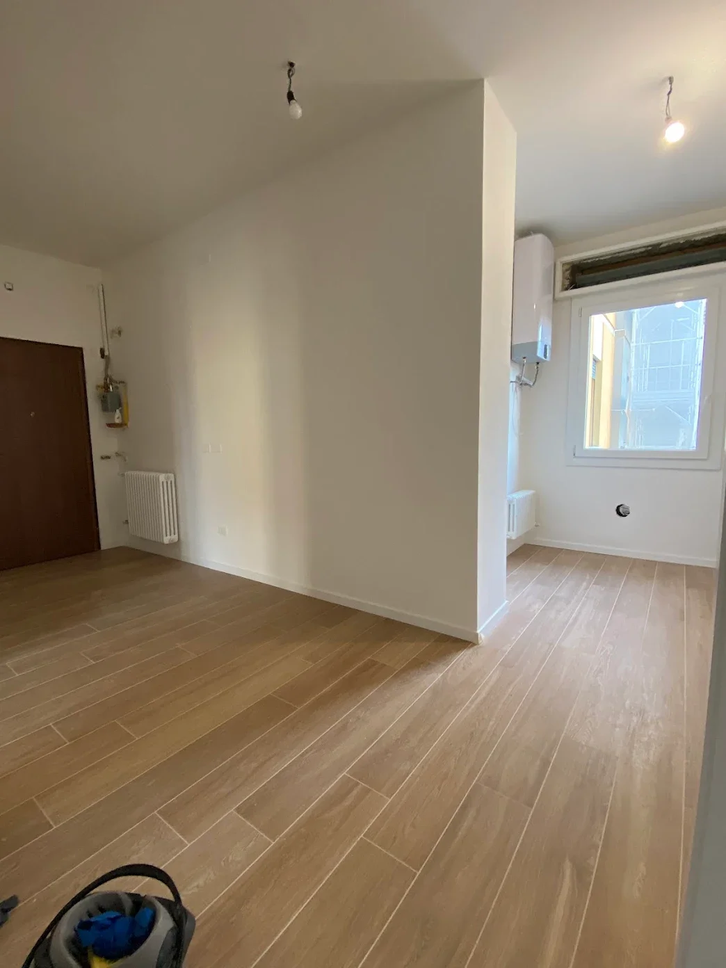 Interno di un appartamento in fase di ristrutturazione, con pareti bianche, pavimento in legno e luci non ancora installate, vista zona ingresso e cucina con finestra.