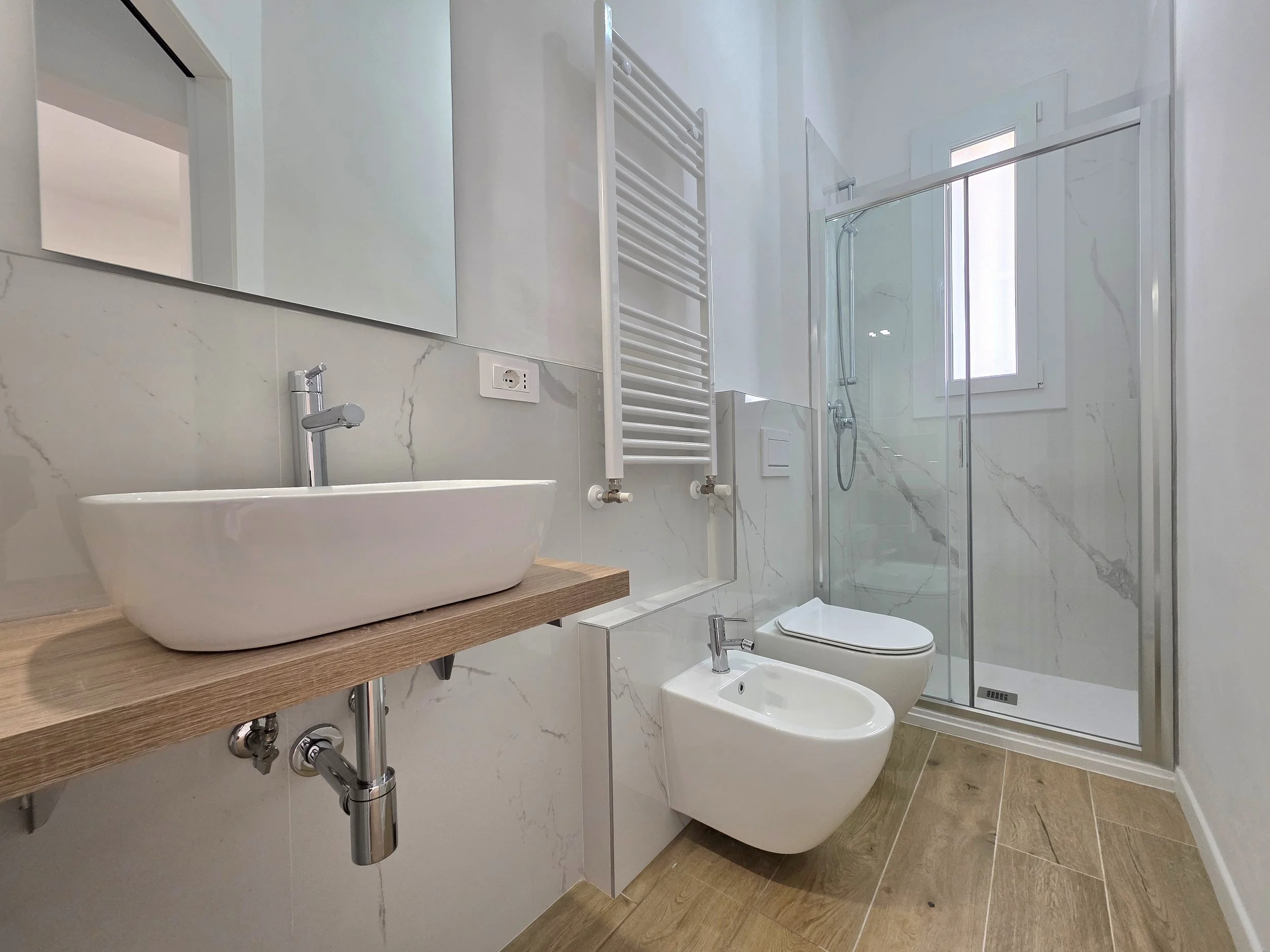 Bagno moderno con lavabo su mensola di legno, specchio grande, termosifone bianco a parete, bidet e WC, doccia con parete di vetro e finestra, pavimento in legno