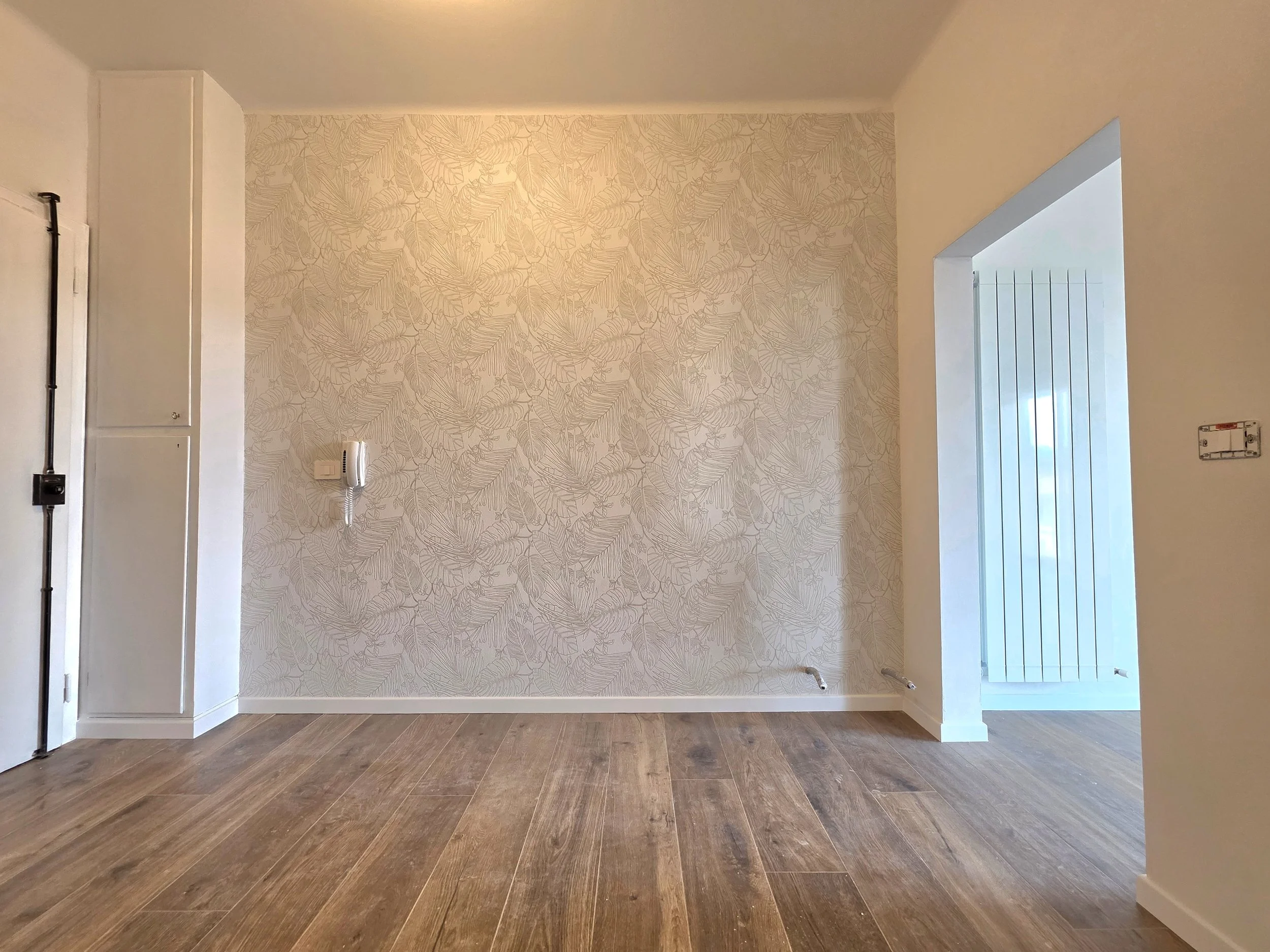 Interno di una stanza con pareti beige, una parete con carta da parati a motivo foglie e un pavimento in legno.