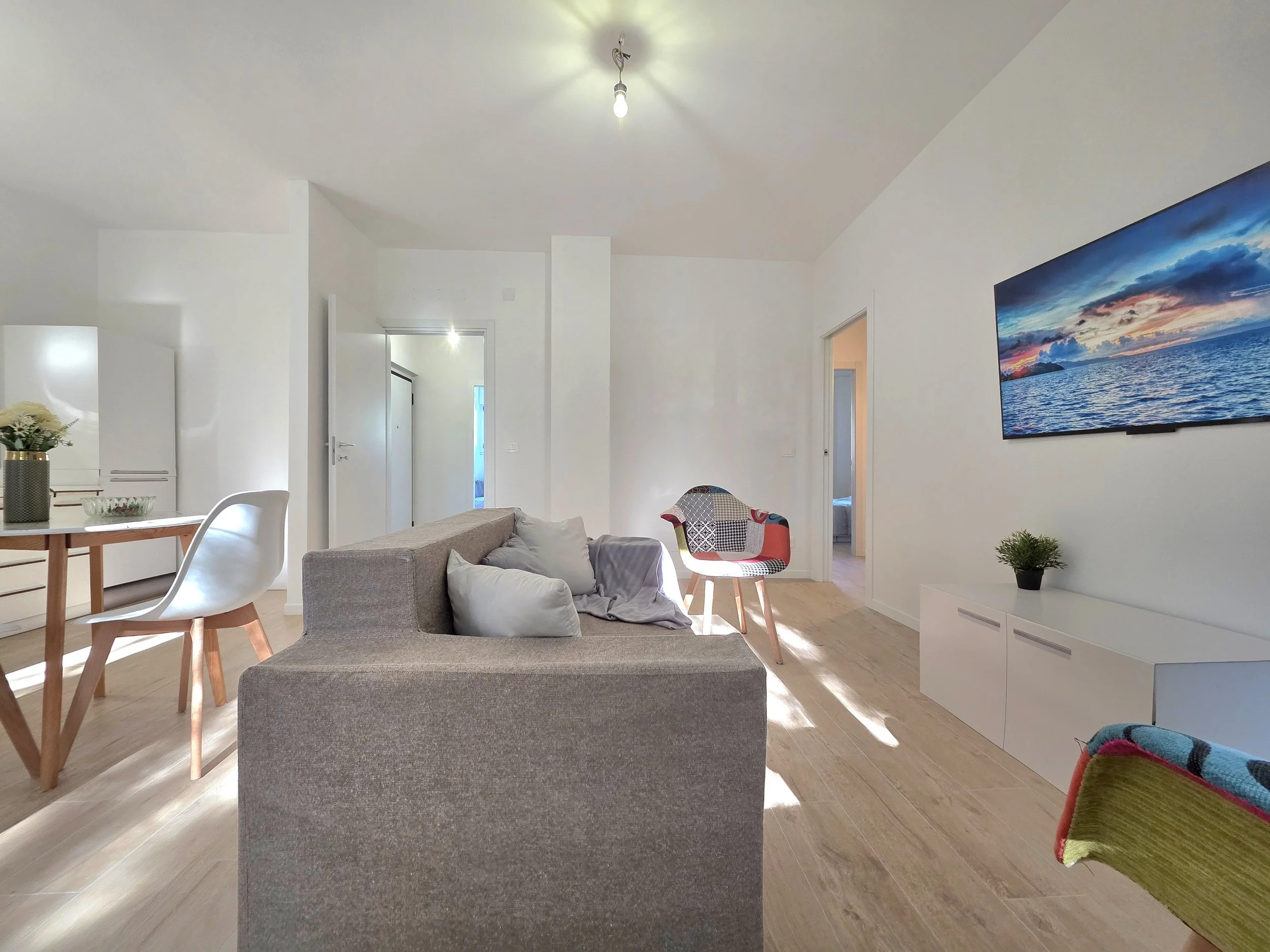 Interno di un soggiorno con pareti bianche, divano grigio, televisore a schermo piatto con immagine di un tramonto sul mare, tavolo in legno con sedie bianche, poltrona colorata con patchwork, pavimento in legno chiaro, luci accese.