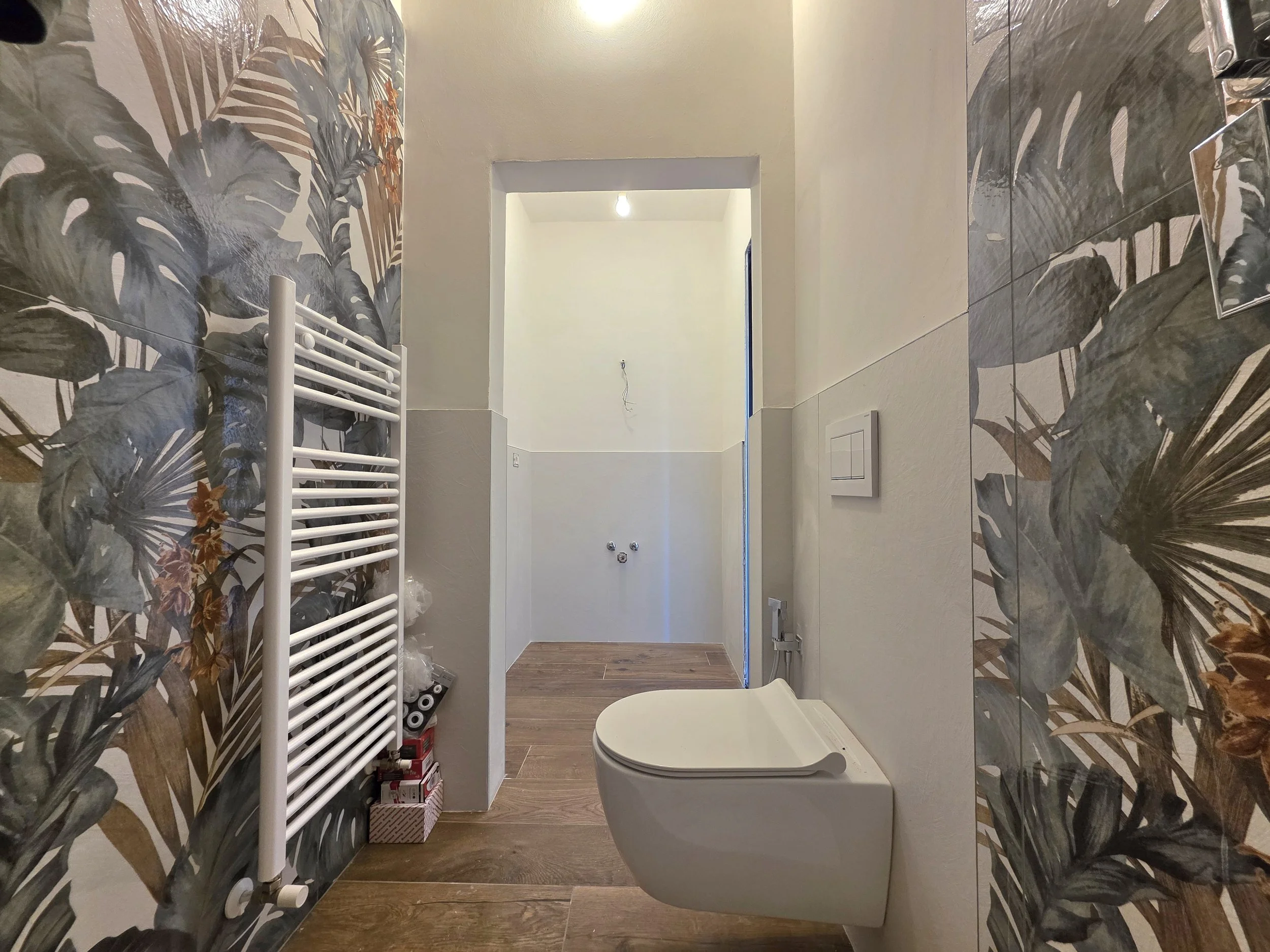 Una stanza da bagno in fase di completamento, con una parete decorata con carta da parati con motivi di grandi foglie, un asciugamano caldario bianco sulla sinistra, e un sanitari appoggiato alla parete sulla destra.