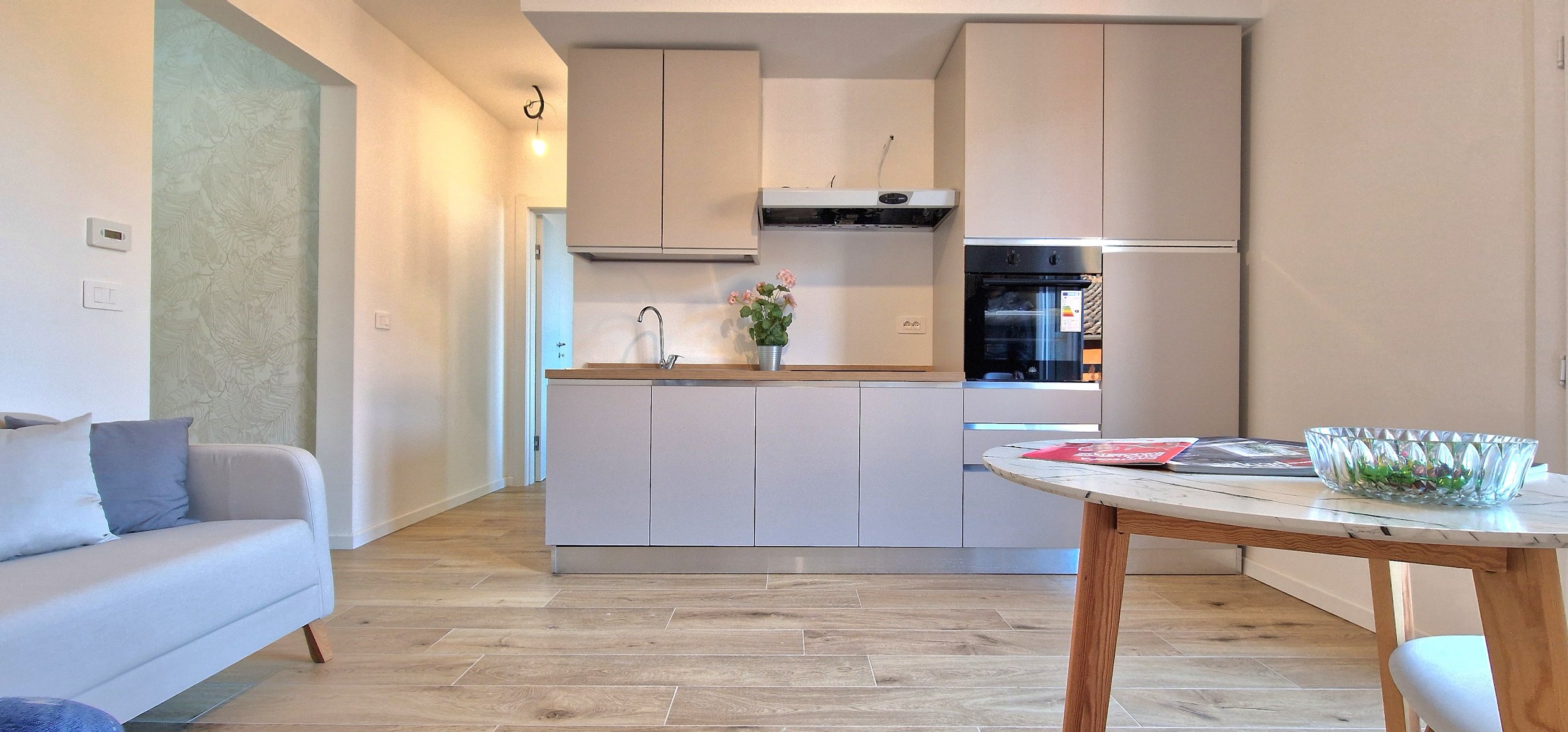 Cucina moderna con armadietti beige, forno integrato, penisola con lavabo e piante, soggiorno con divano e tavolo in legno con ciotola decorativa.